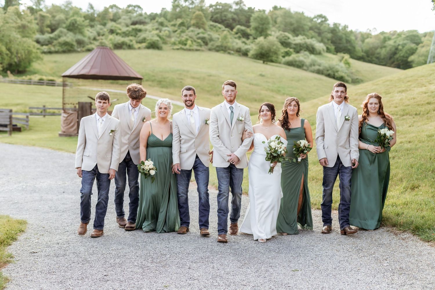 Wedding party wearing sage green and beige attire poses together on gravel path in countryside setting.