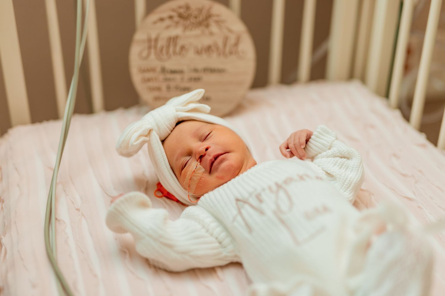 A sleeping newborn baby in white clothing lies in a crib with a wooden 'Hello World' sign visible above.