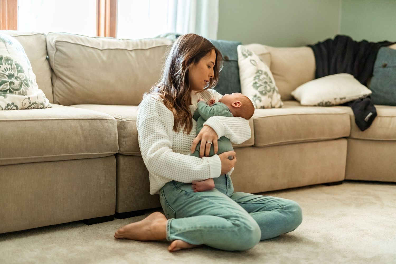 Someone feeds a newborn baby while sitting on the floor by a beige couch in a living room.