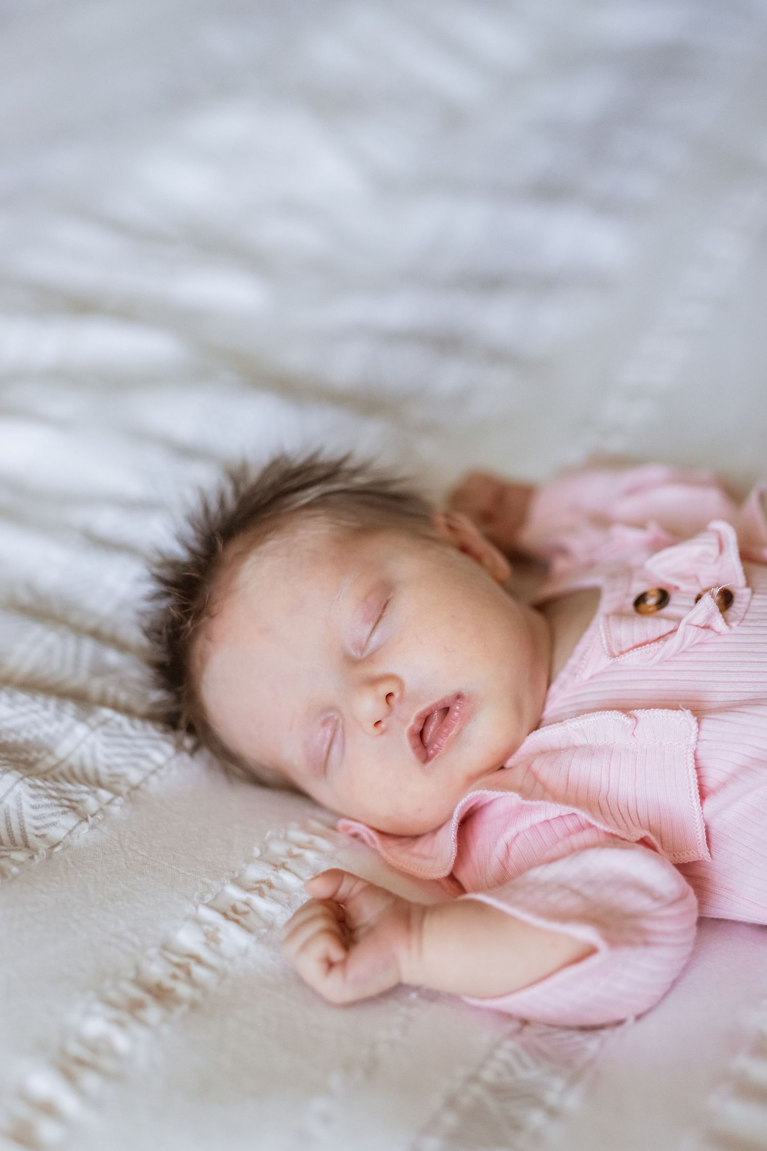 A peaceful newborn baby sleeping in a pink onesie on a soft white blanket.