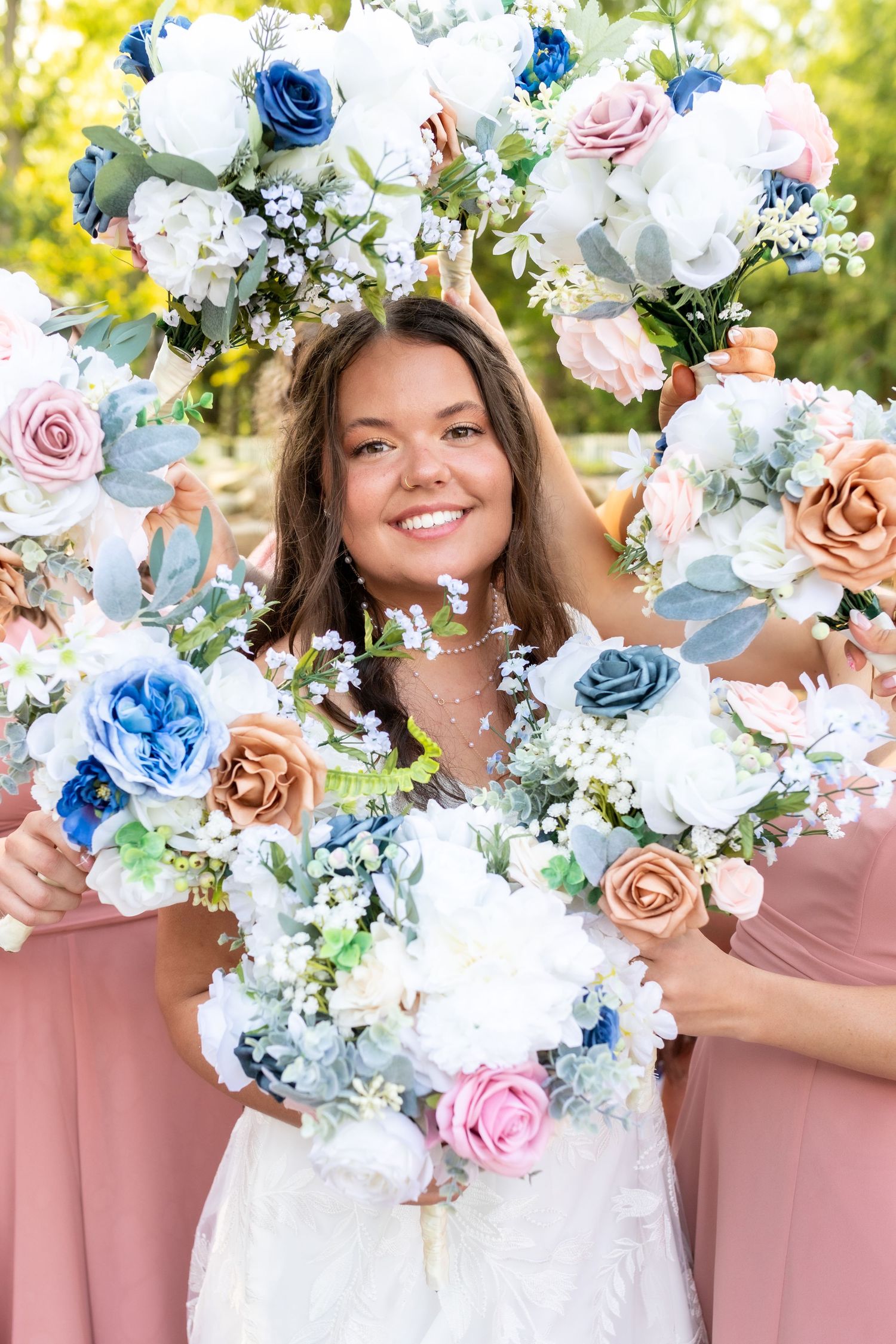 Smiling bride surrounded by bridesmaids holding blue, pink, and white floral bouquets outdoors.