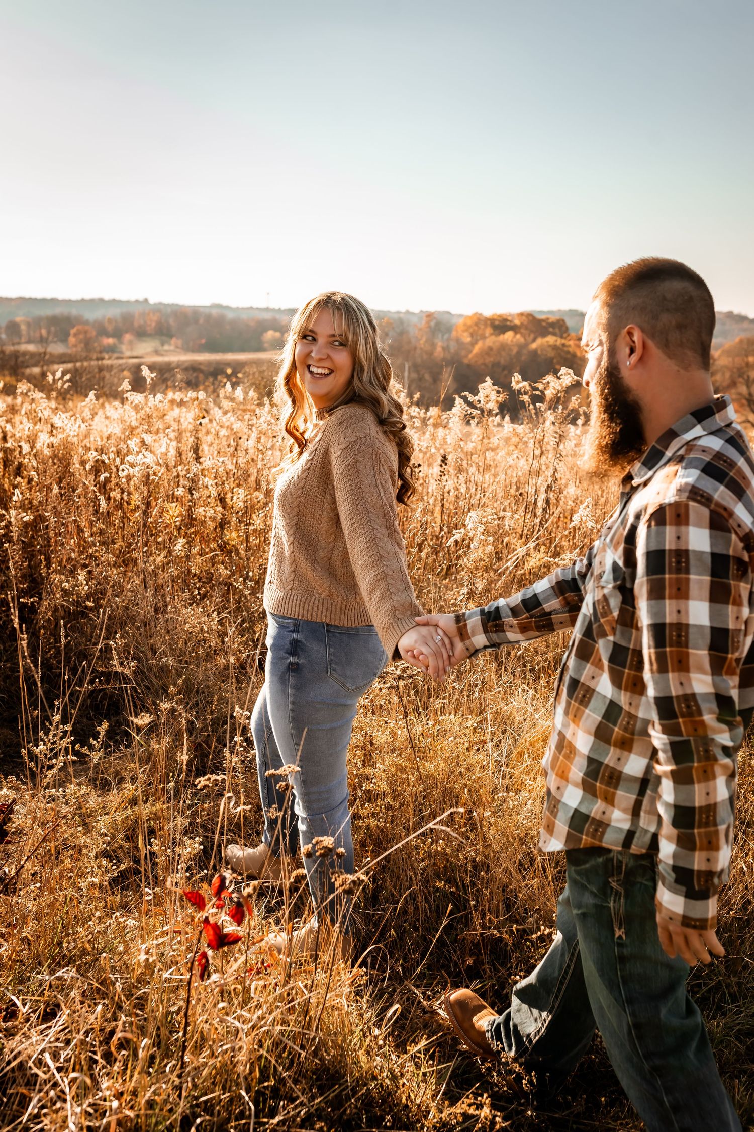 A couple holding hands in a golden sunlit meadow during autumn wearing casual attire.
