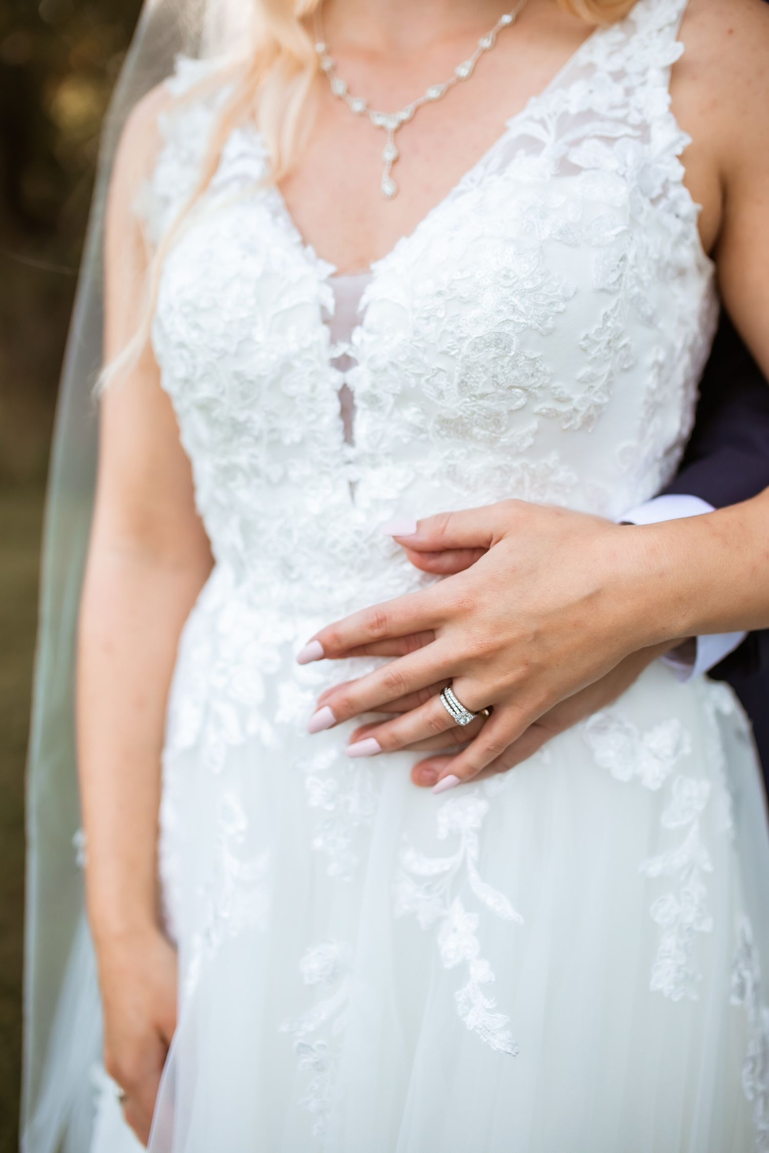 Close-up detail of white lace wedding dress with delicate necklace and engagement ring displayed on hand.
