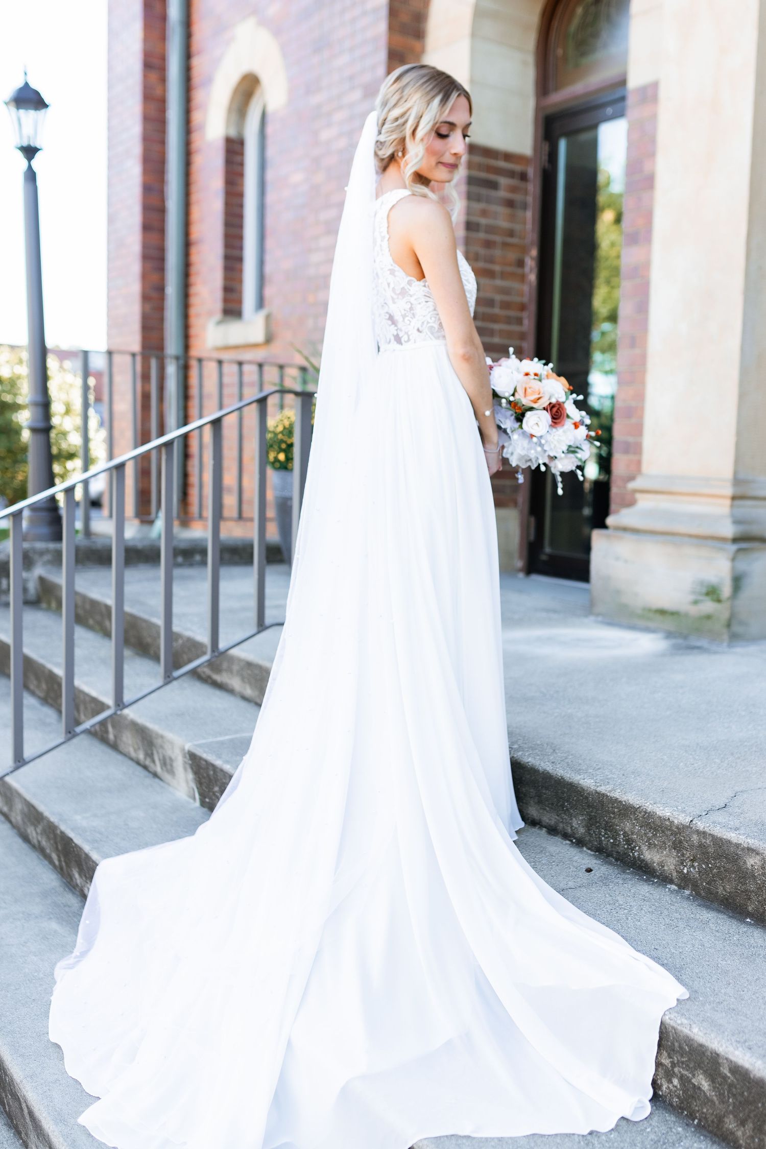 A bride in an elegant white flowing gown with train stands on church steps holding a white and blush floral bouquet.