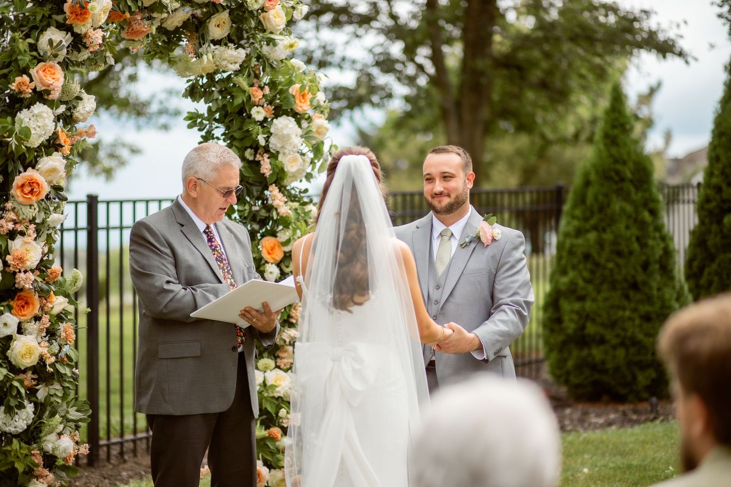 Outdoor wedding ceremony with groom and officiant at floral arch with peach and white flowers.