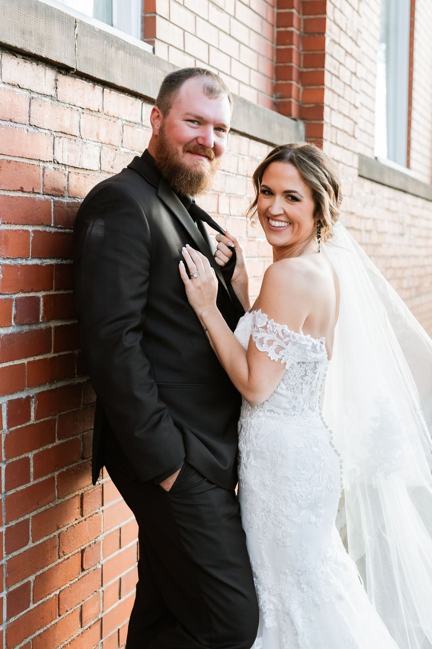 A happy couple poses together against a brick wall on their wedding day, wearing a white lace gown and black suit.