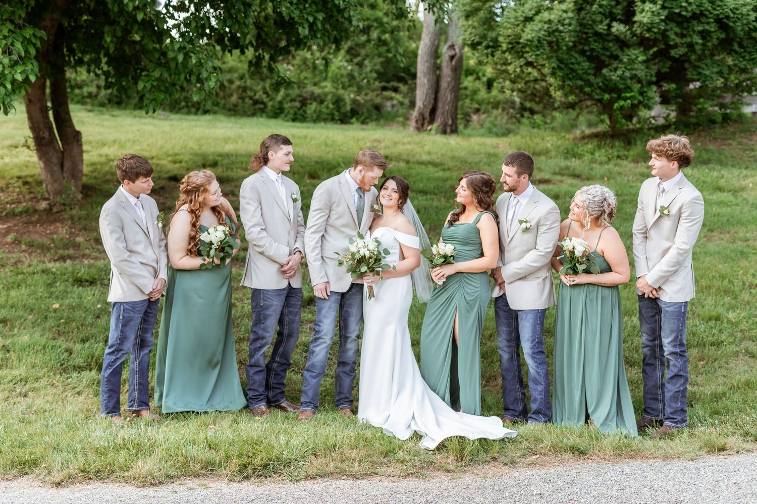 Wedding party wearing sage green dresses and gray suits poses together on grassy path during outdoor ceremony.