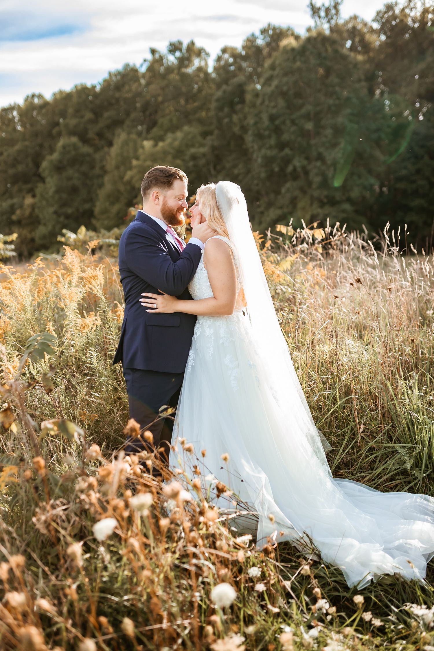 A romantic couple embraces in a sunlit meadow with tall grass and wildflowers during their outdoor wedding celebration.
