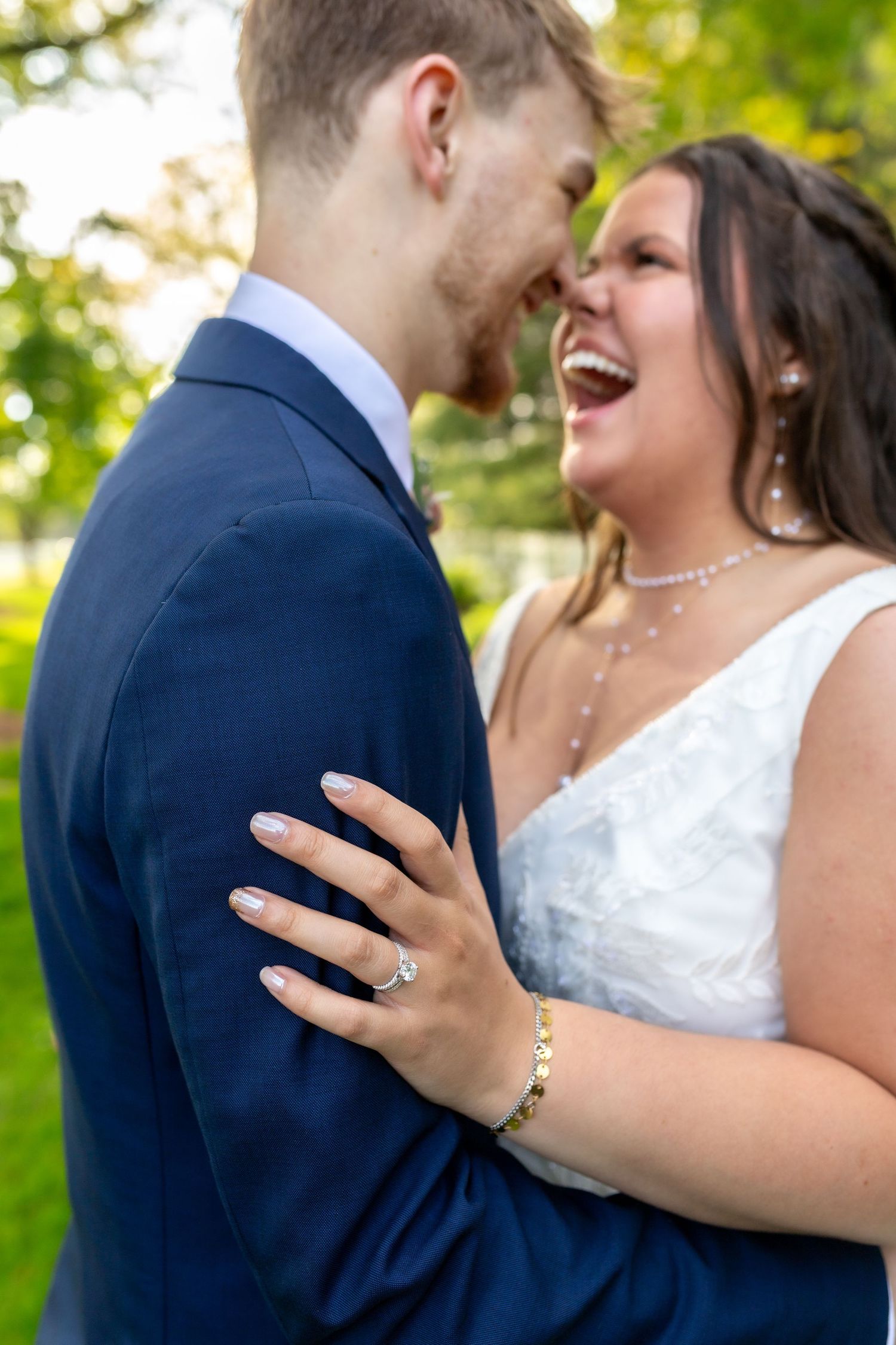 Laughing bride in white dress shows off engagement ring while embracing groom in navy suit outdoors.