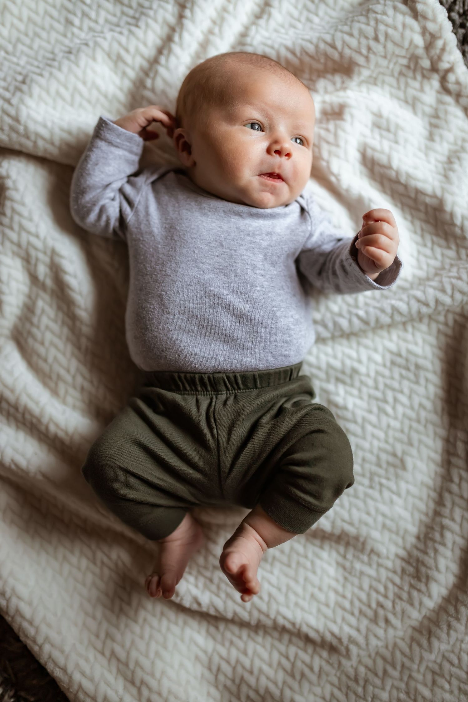Sweet baby wearing a grey sweater and olive-colored pants lying on a white textured blanket.