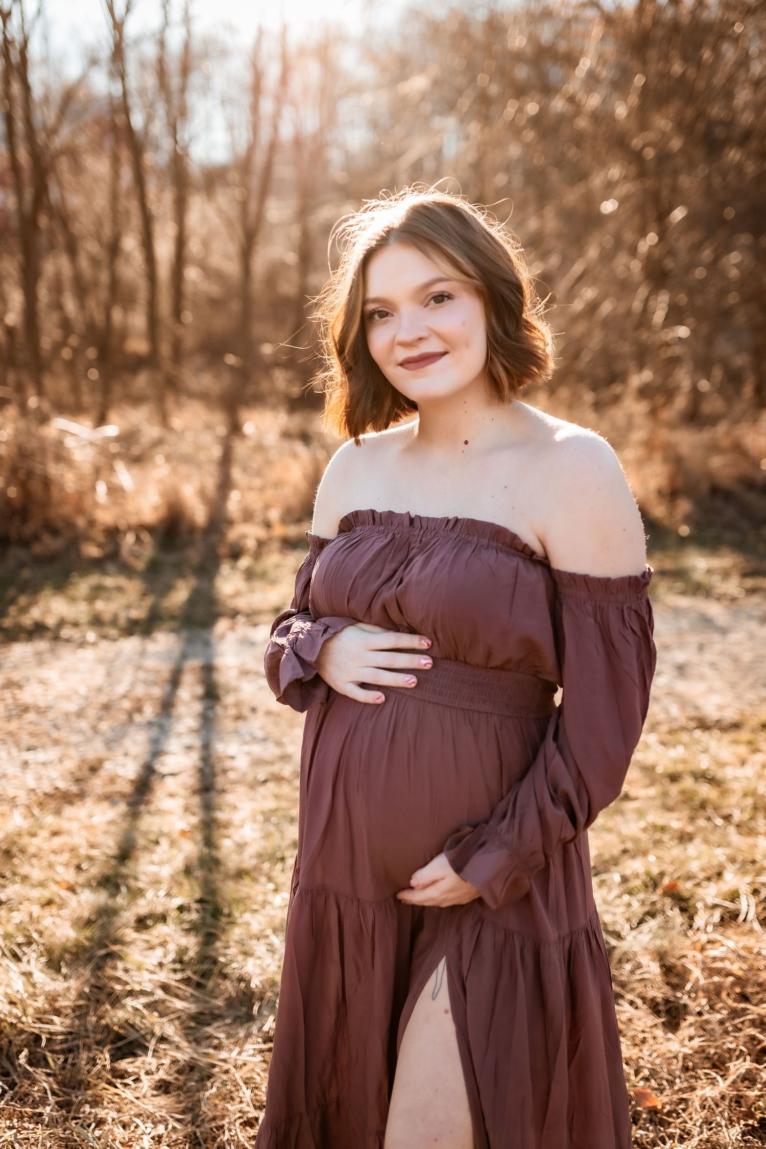 A pregnant woman in a burgundy off-shoulder dress poses outdoors during golden hour in late summer.