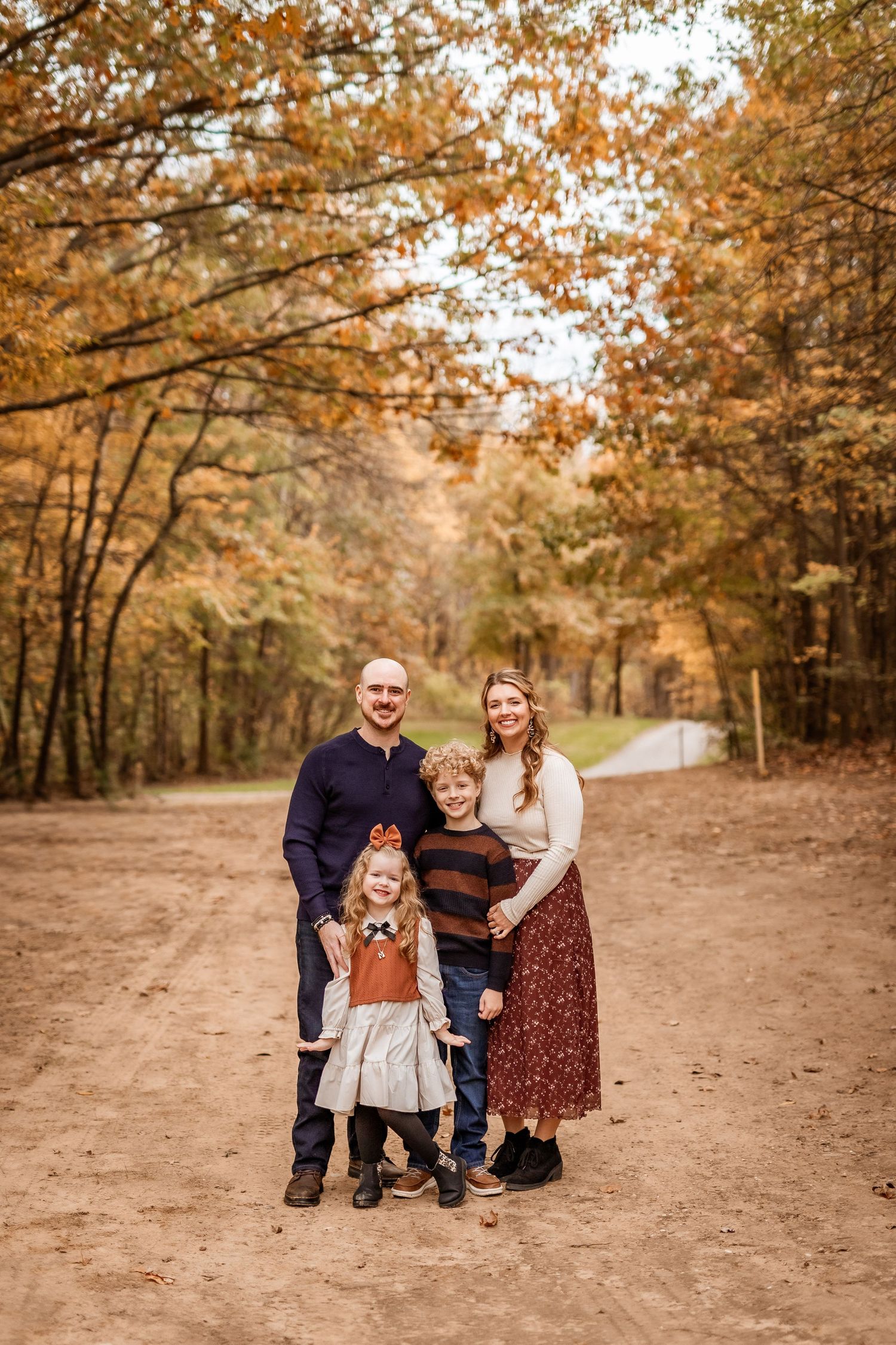 A family poses together on a dirt path surrounded by autumn trees with golden leaves in a forest setting.