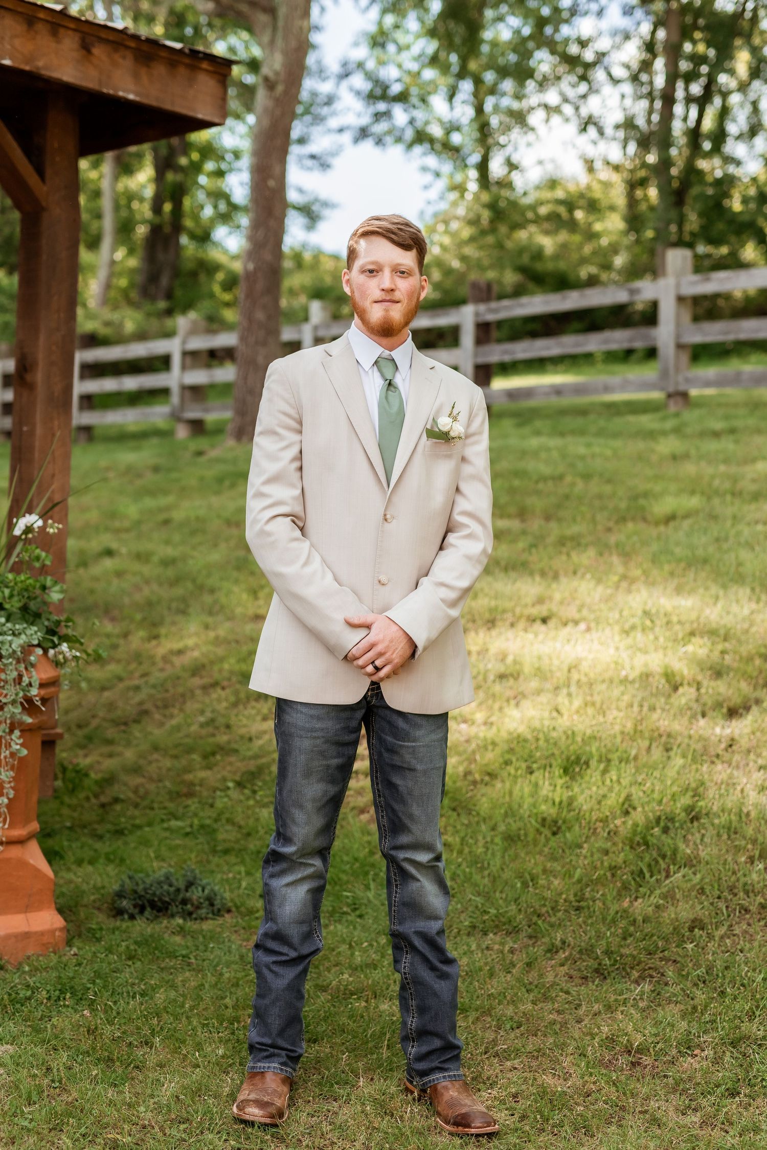 Someone wearing a beige blazer with a mint green tie stands in a rustic outdoor setting with wooden fence in background.