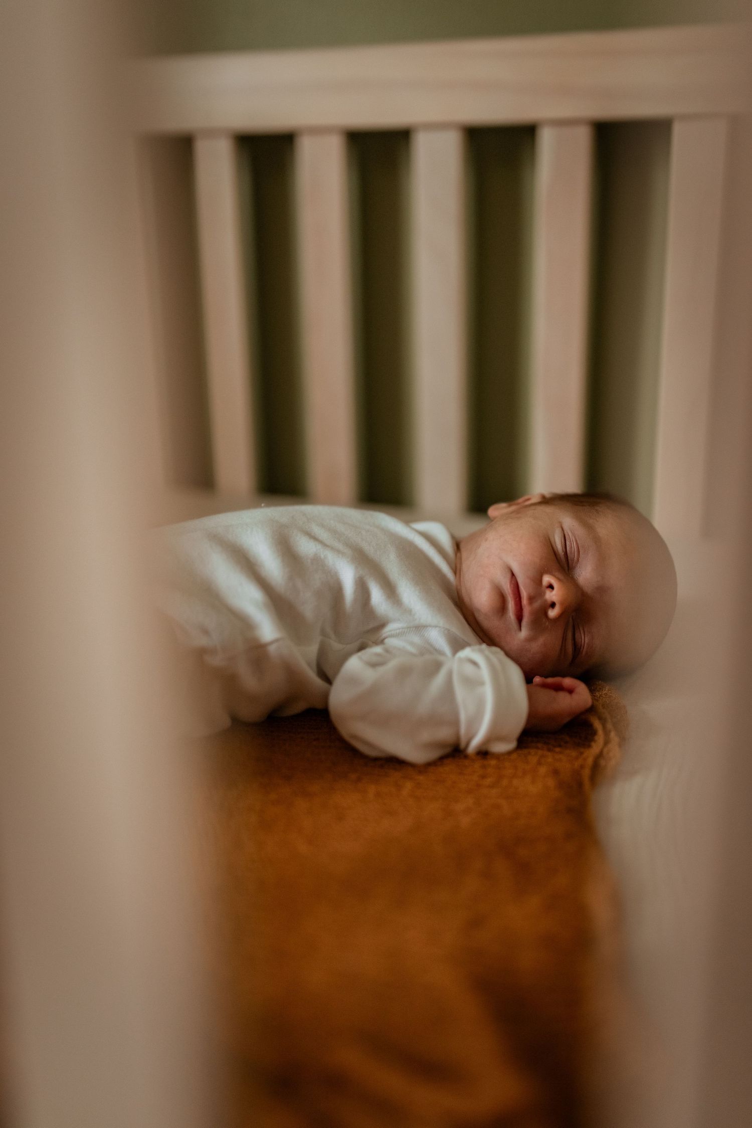 A sleeping baby wearing white sleepwear rests peacefully on brown bedding, viewed through crib slats in a nursery setting.