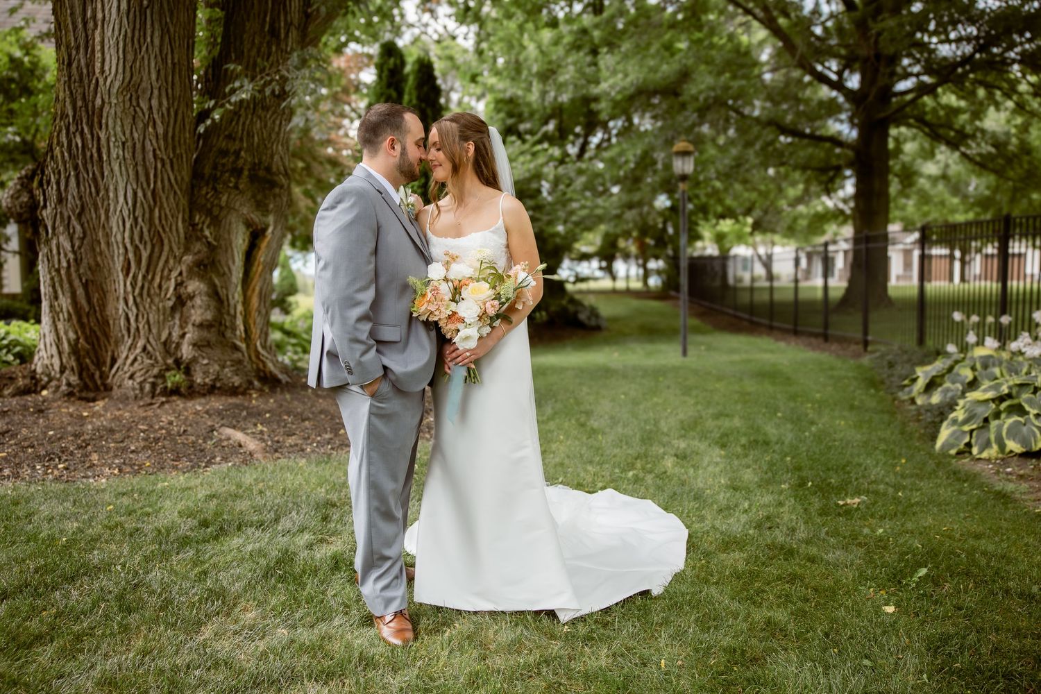 Bride and groom share a romantic moment outdoors, bride holding a pastel bouquet, groom in gray suit, lush green garden backdrop.
