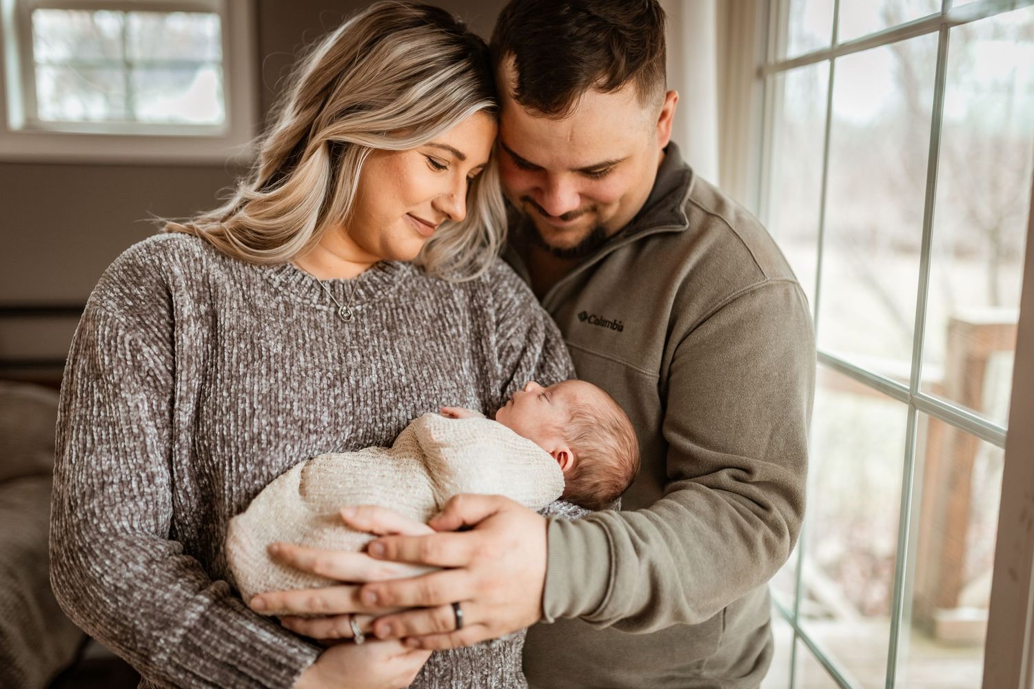 A couple in cozy sweaters tenderly holds their newborn baby near a bright window in an intimate indoor setting.