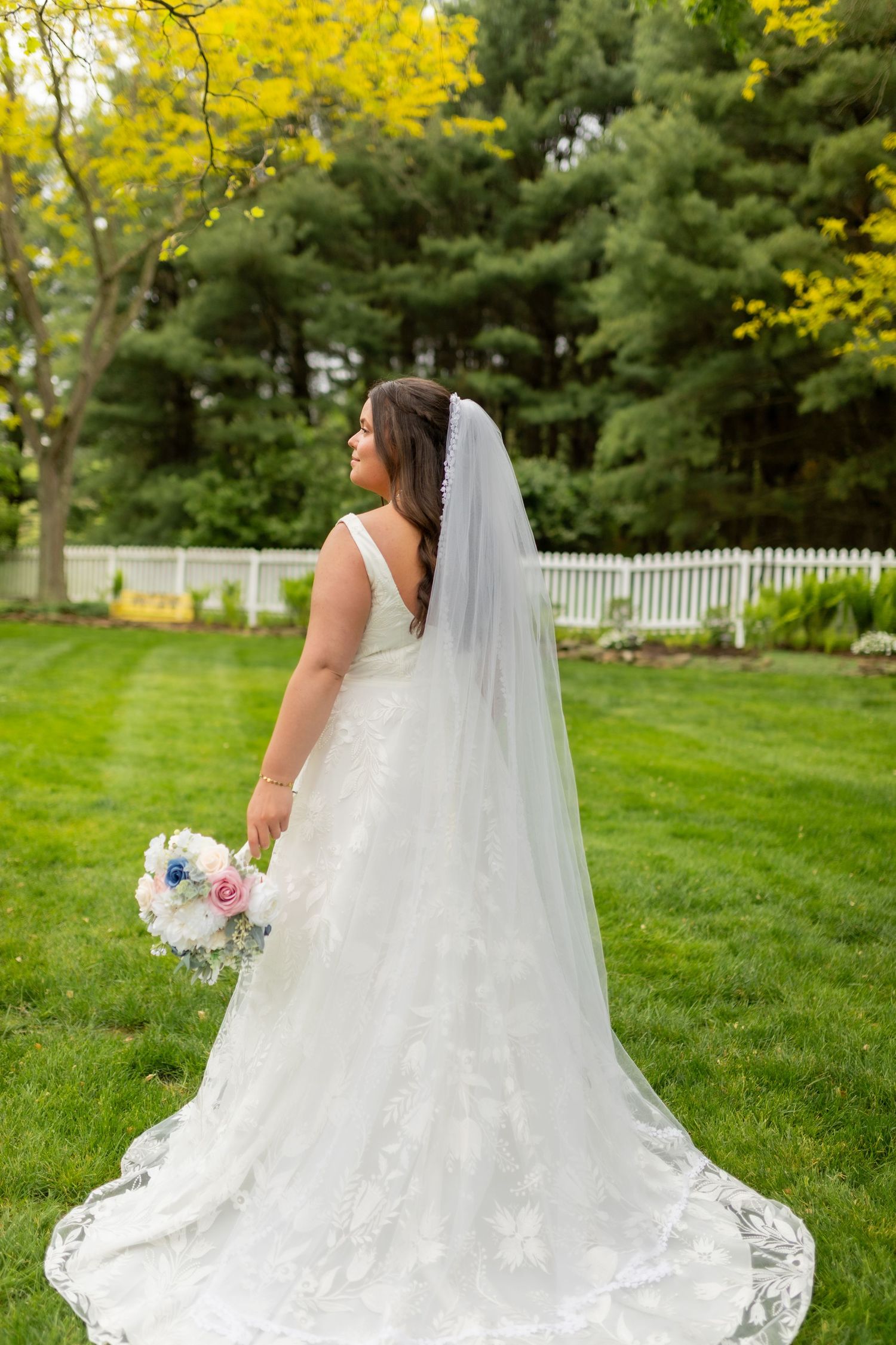 Bride in lace wedding gown with veil holding bouquet standing in lush green garden.