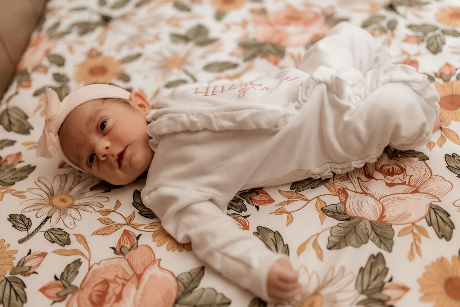 Newborn baby wearing white sleepwear rests on floral patterned bedding with soft peach and green tones.