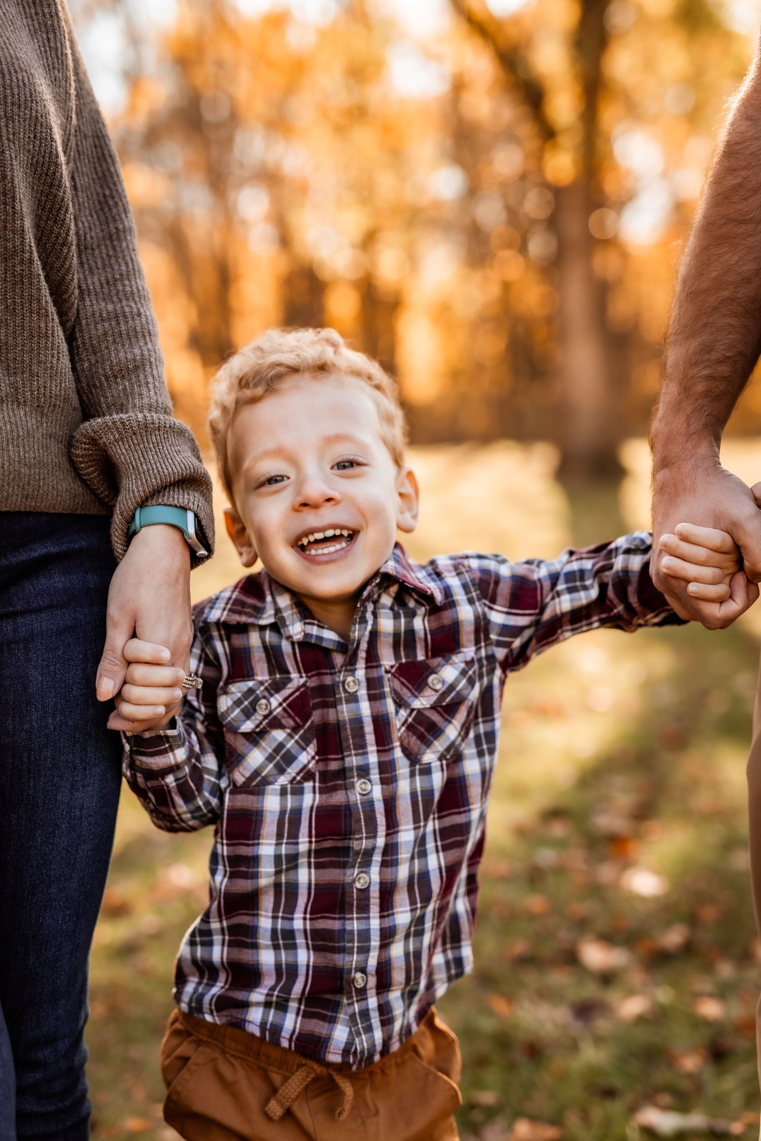 Happy child wearing plaid shirt holds hands with parents while playing outdoors on autumn day with golden trees in background.