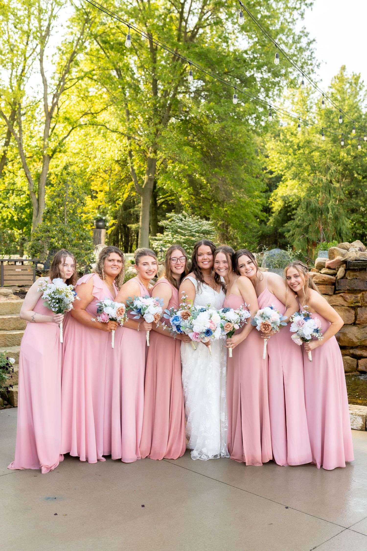 Bride in white gown poses with six bridesmaids in pink dresses holding floral bouquets outdoors.