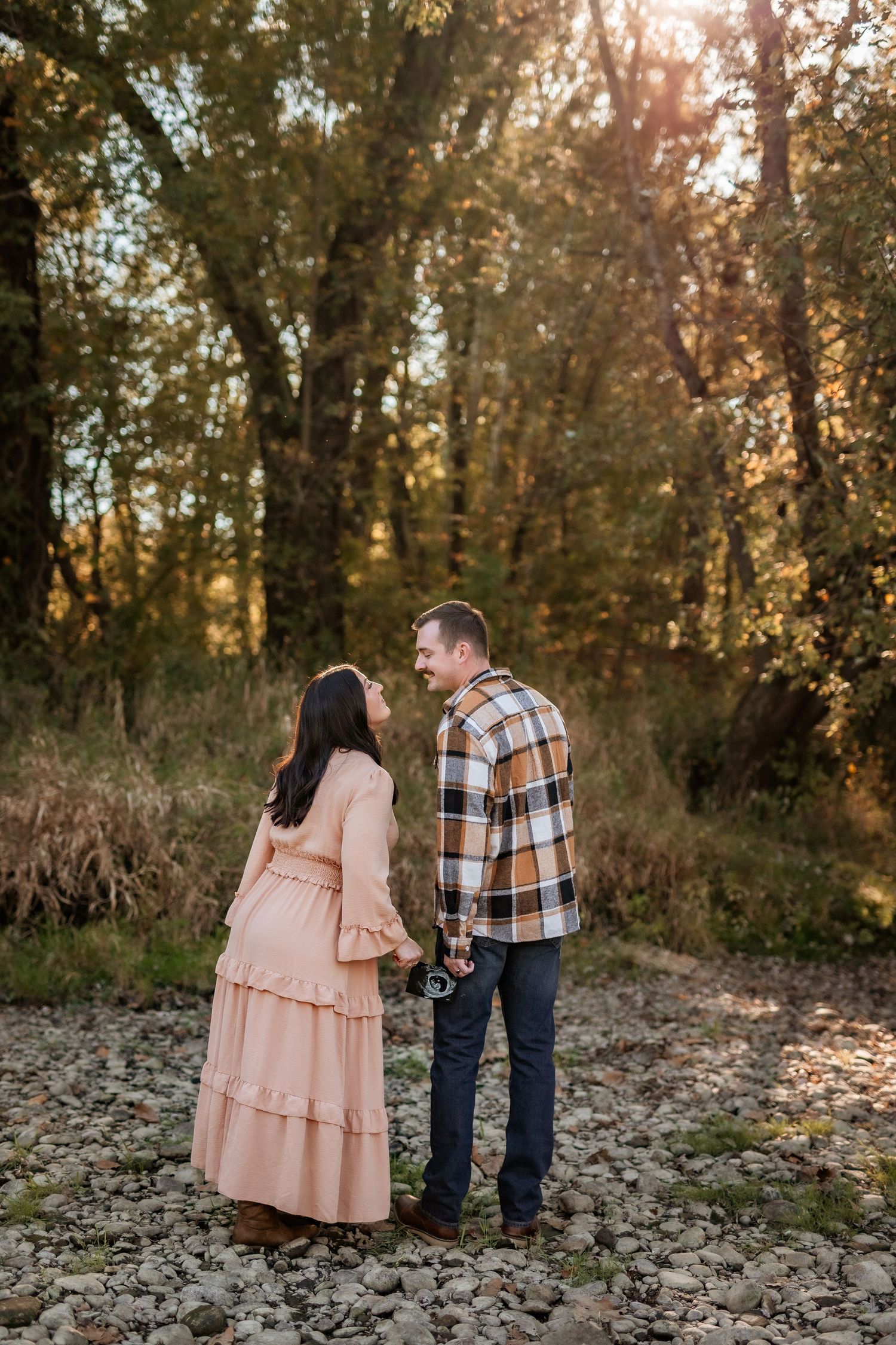 Pregnant couple standing together on rocky ground in golden autumn forest, woman in pink dress, man in plaid shirt.