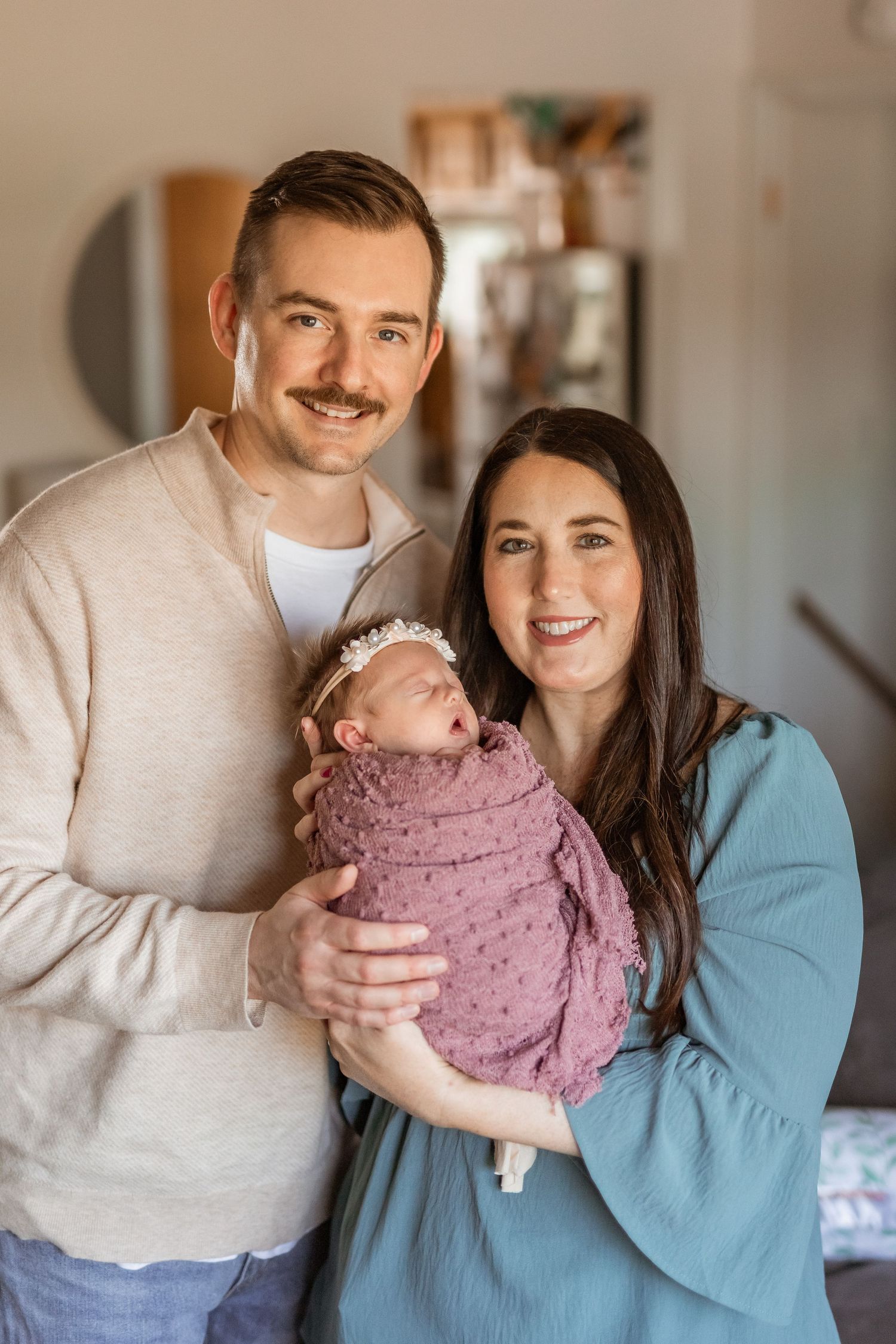 A smiling couple holds their swaddled newborn baby girl wearing a floral headband in a warm indoor setting.
