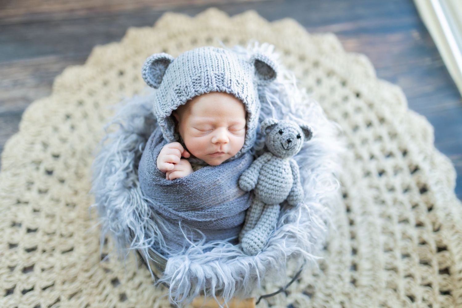 Sleeping newborn baby in gray bear knit hat and wrap, holding teddy bear in wooden bowl on crochet blanket.
