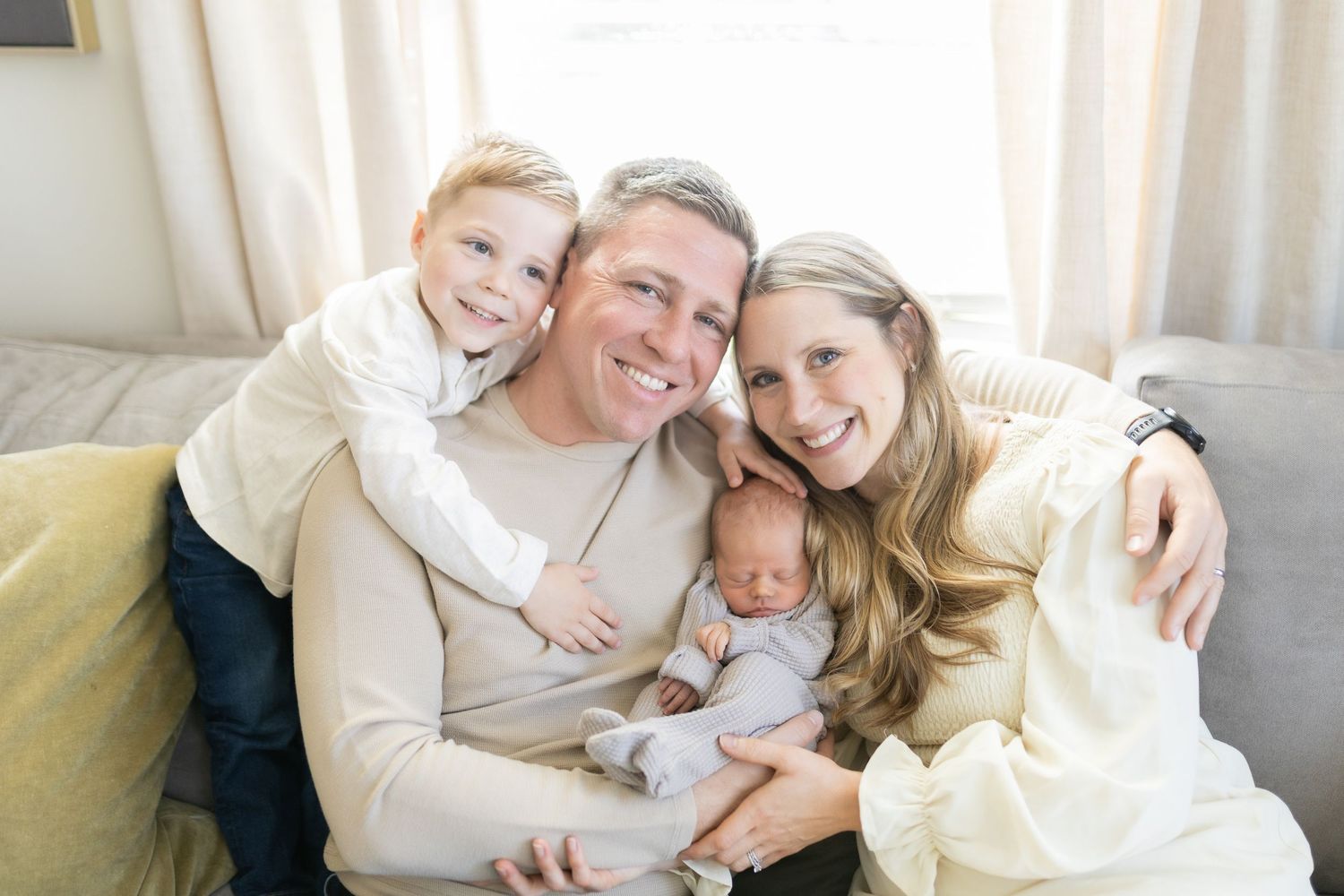 Happy family of four posing on a couch, parents smiling with toddler son and newborn baby in a bright living room.