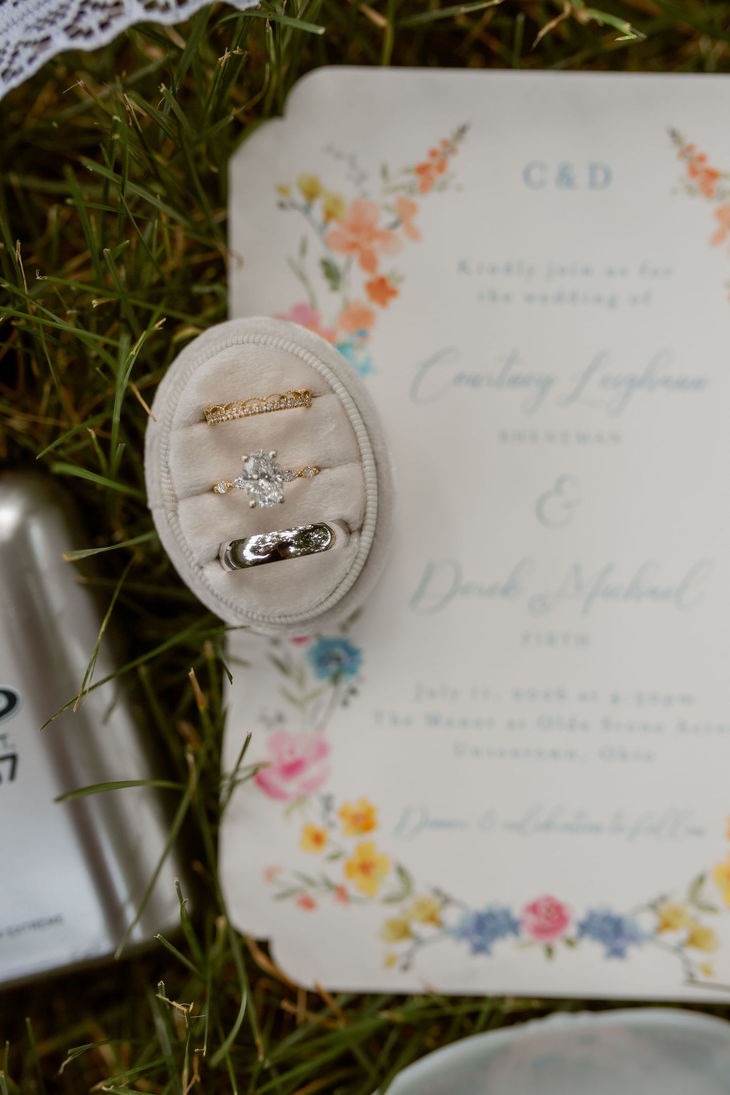 Wedding rings displayed on velvet box beside floral invitation for Courtney and Derek's ceremony in Hutchinson, Ohio.