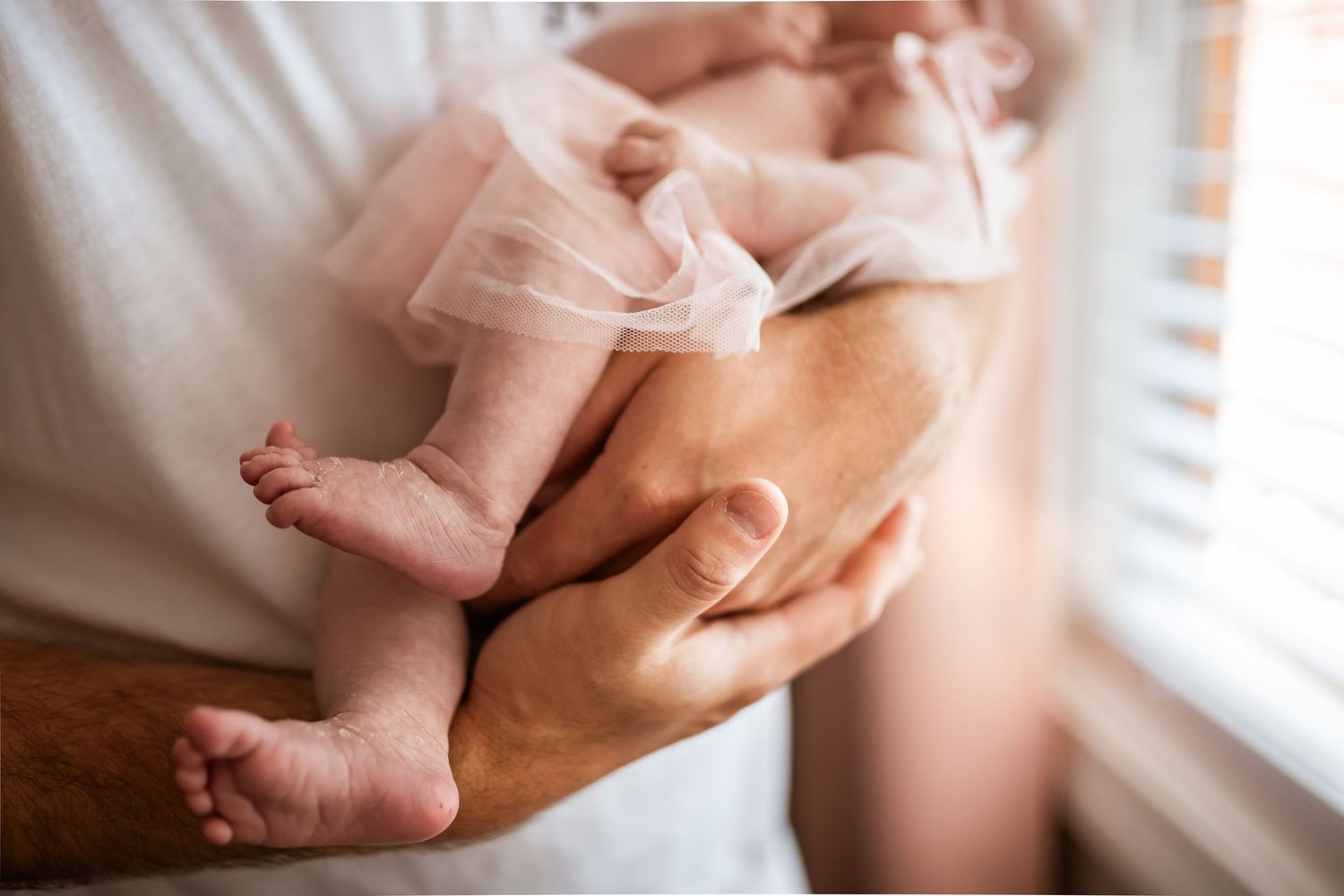 Close-up view of newborn baby feet being gently held by parents' hands near a window with soft natural light.