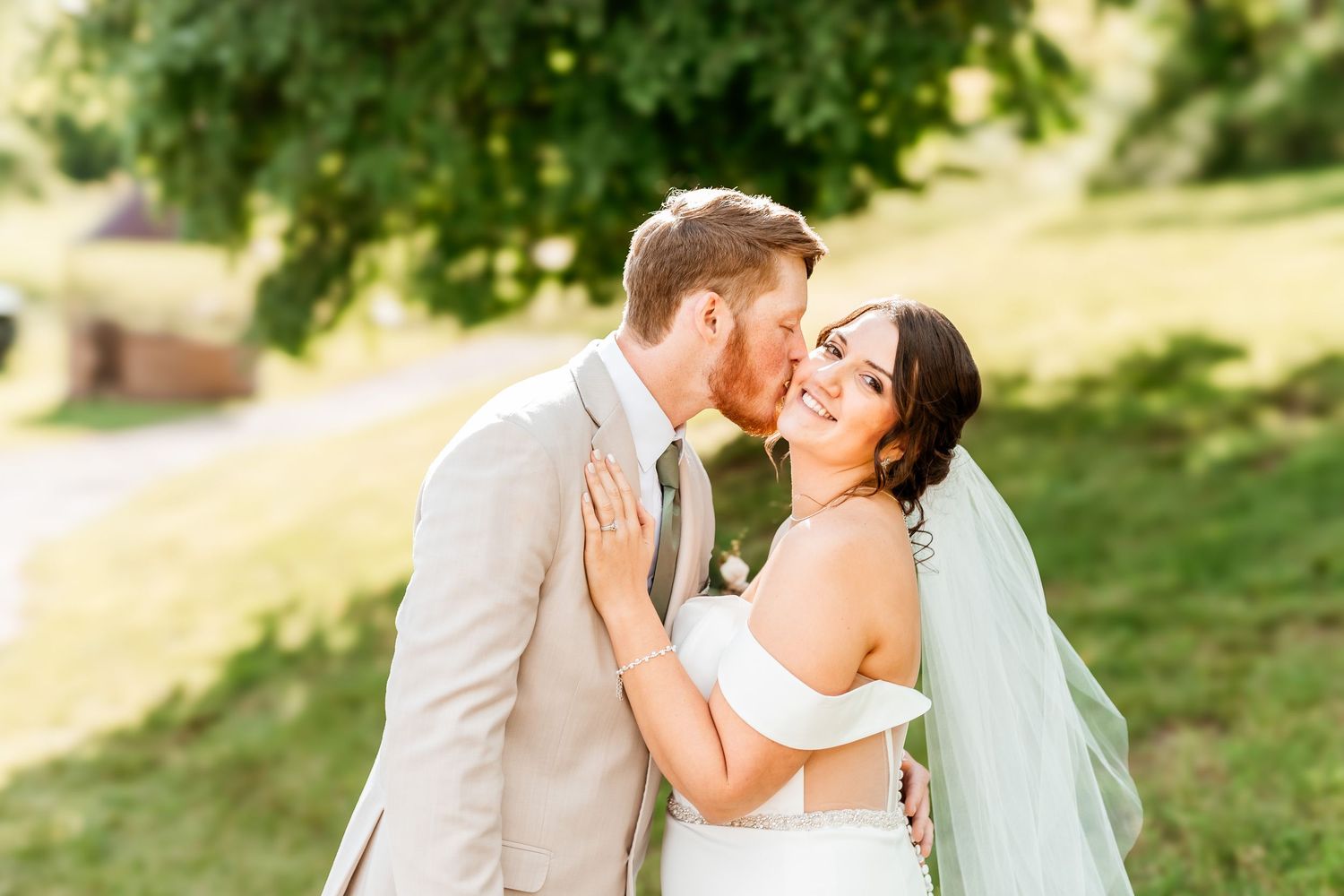 A couple in wedding attire sharing a romantic moment outdoors in a sunny garden setting with a flowing veil.
