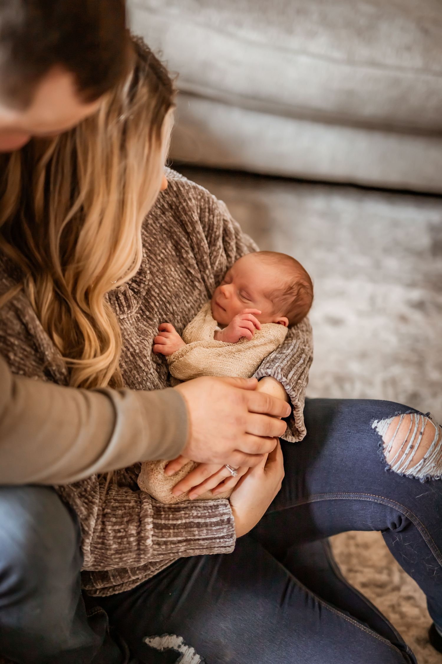 A sleeping newborn baby wrapped in a knit blanket rests in arms wearing a gray sweater and distressed jeans.