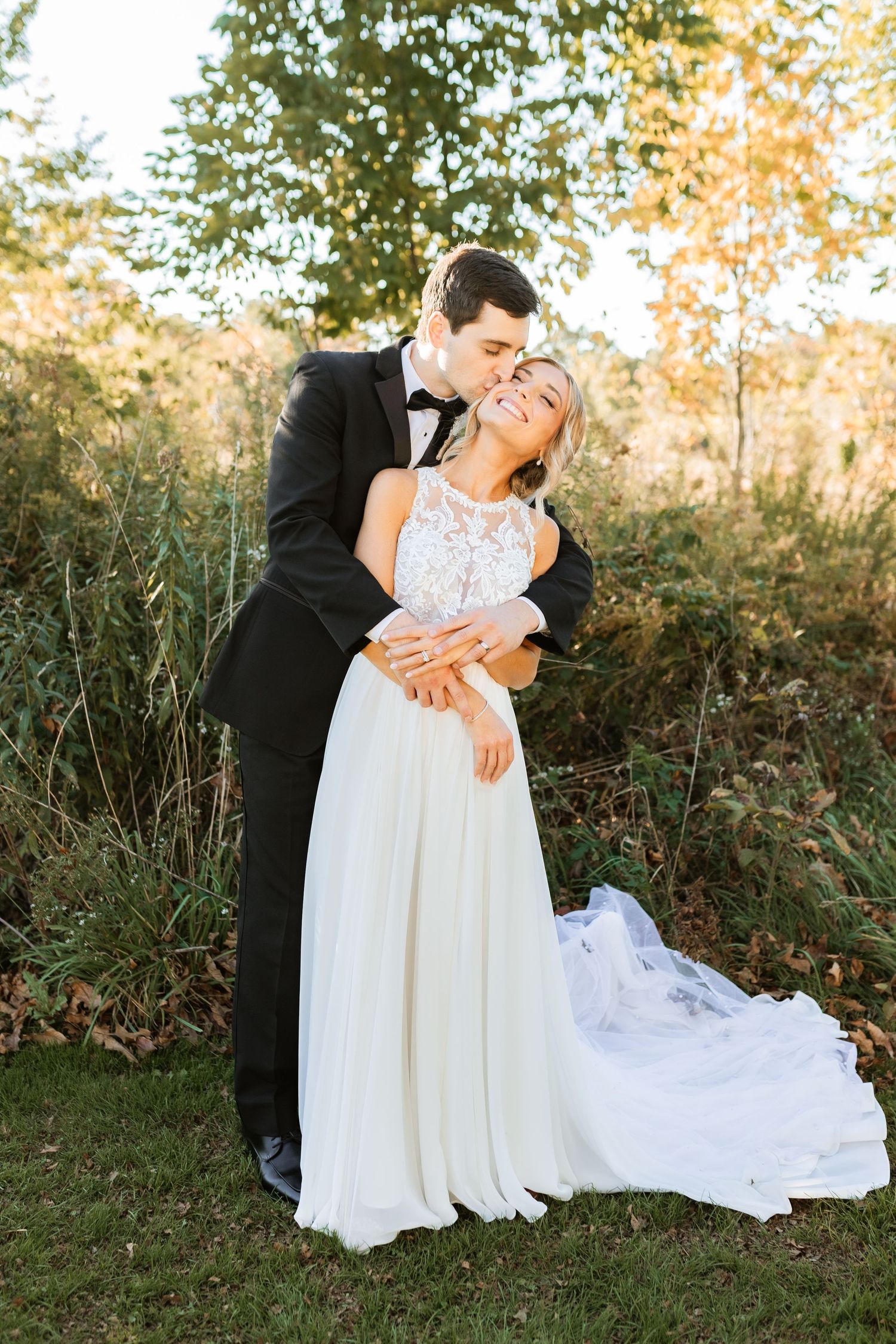 A joyful wedding couple embraces outdoors during golden hour with a beautiful flowing white dress and classic tuxedo.
