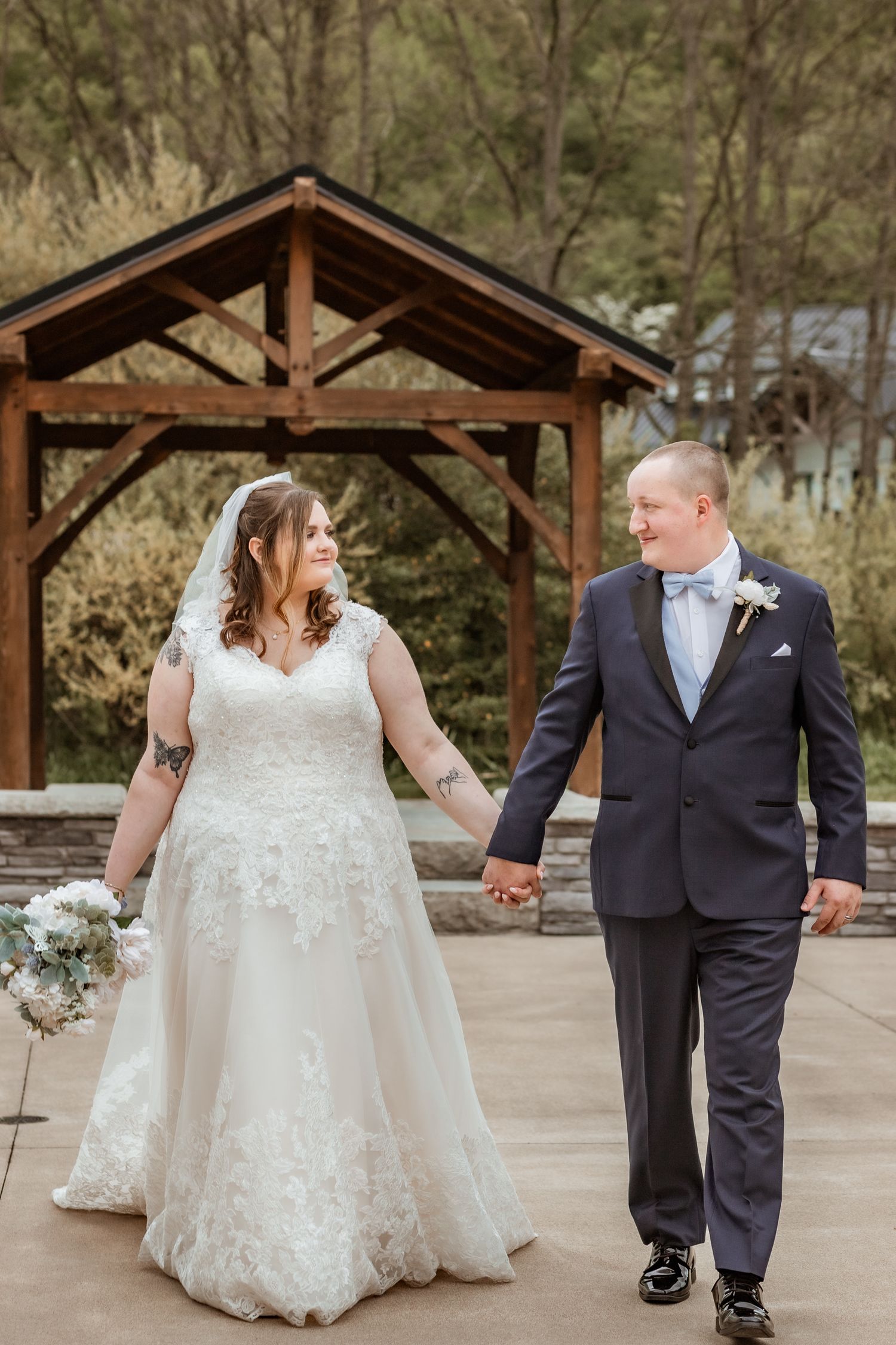 Wedding couple holding hands under a wooden gazebo, bride in lace gown and groom in formal suit.