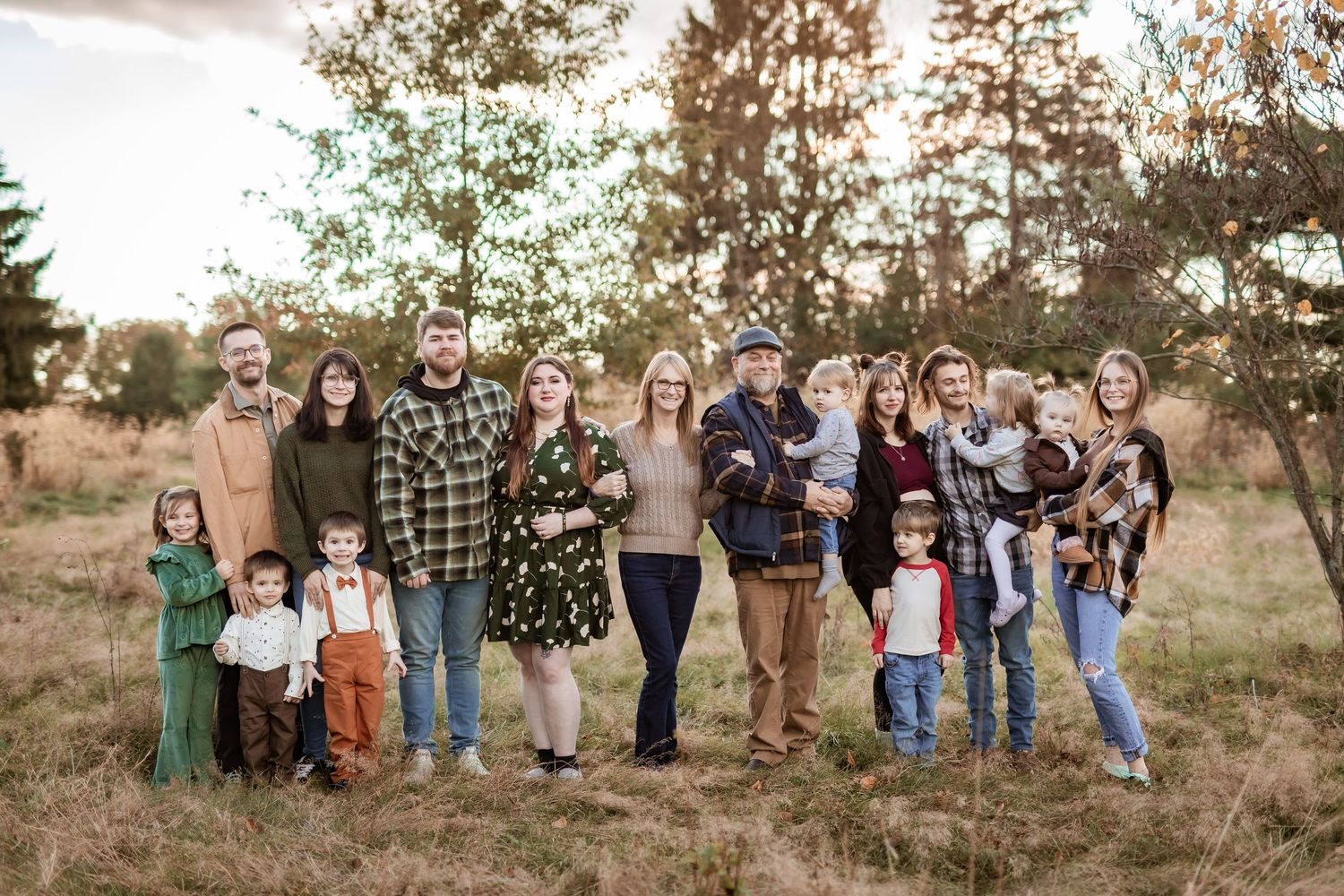Large family group portrait outdoors in a field during golden hour, wearing coordinated fall outfits with trees in background.