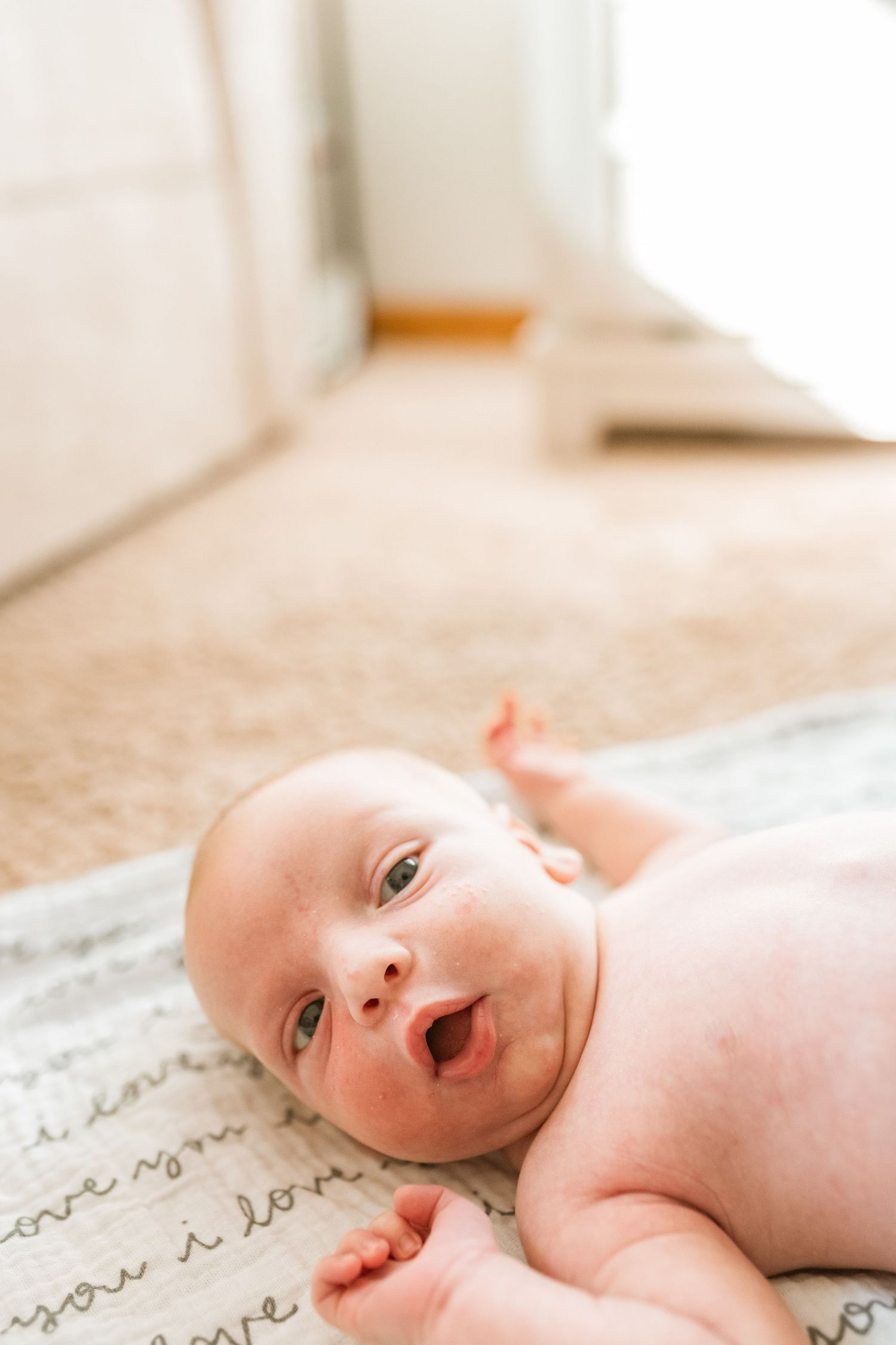 A sweet baby lies on a textured blanket with handwritten text, looking up with bright eyes and an expressive face.