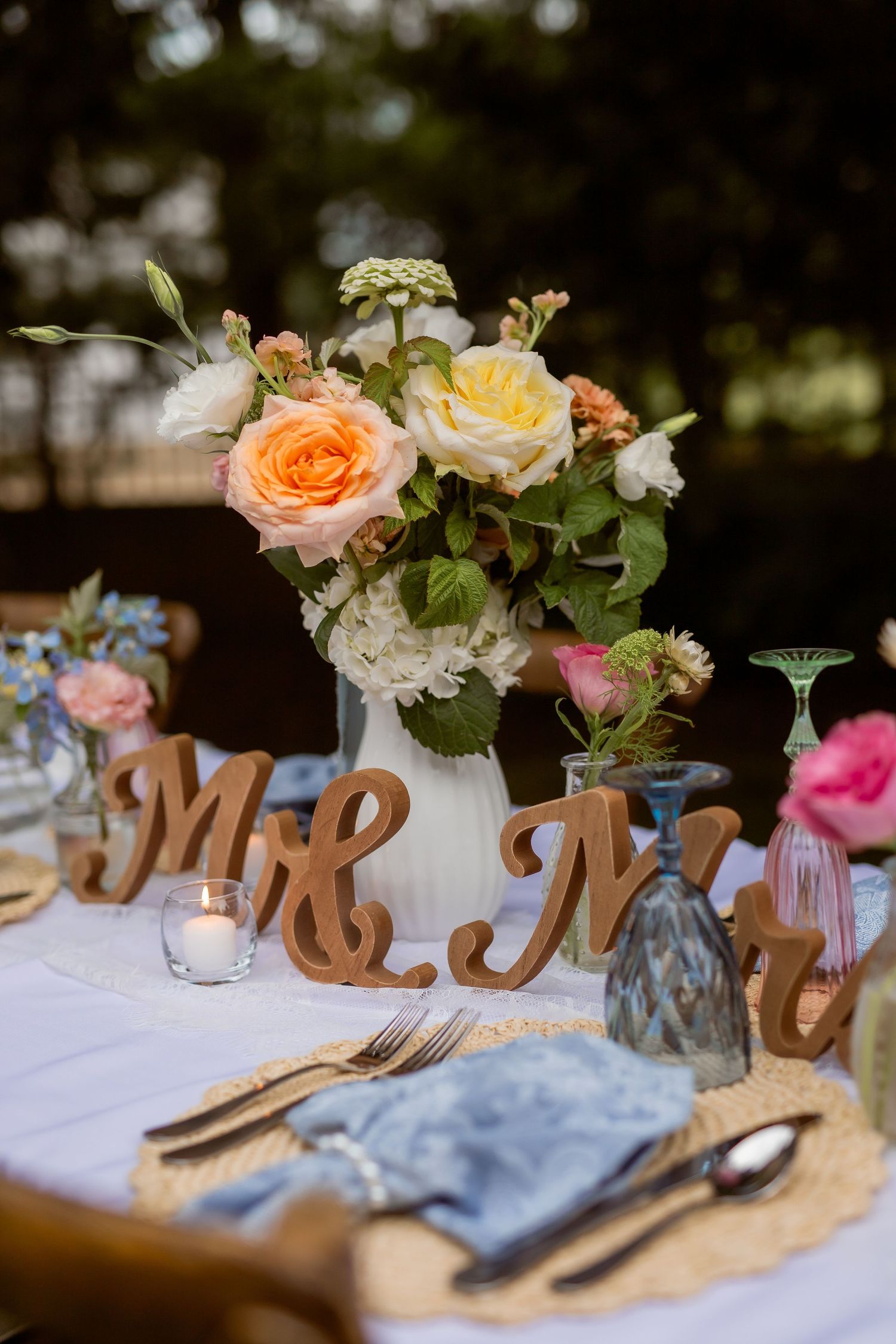 Elegant wedding table setting with Mr. & Mrs. wooden signs, floral centerpiece of peach and yellow roses, candles, and blue napkins.