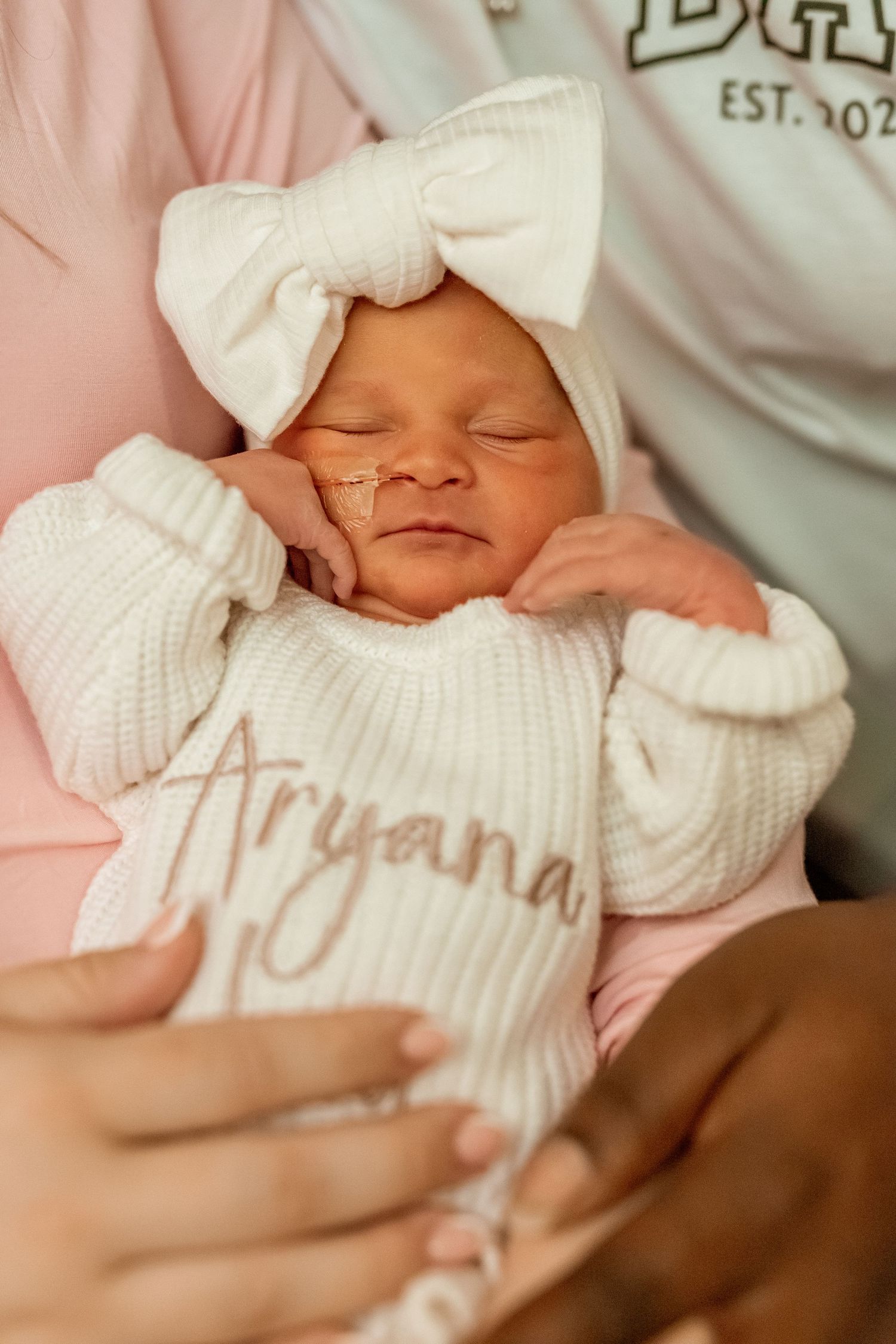 A peaceful sleeping newborn wearing a white knit sweater and matching bow headband rests in soft pink bedding.