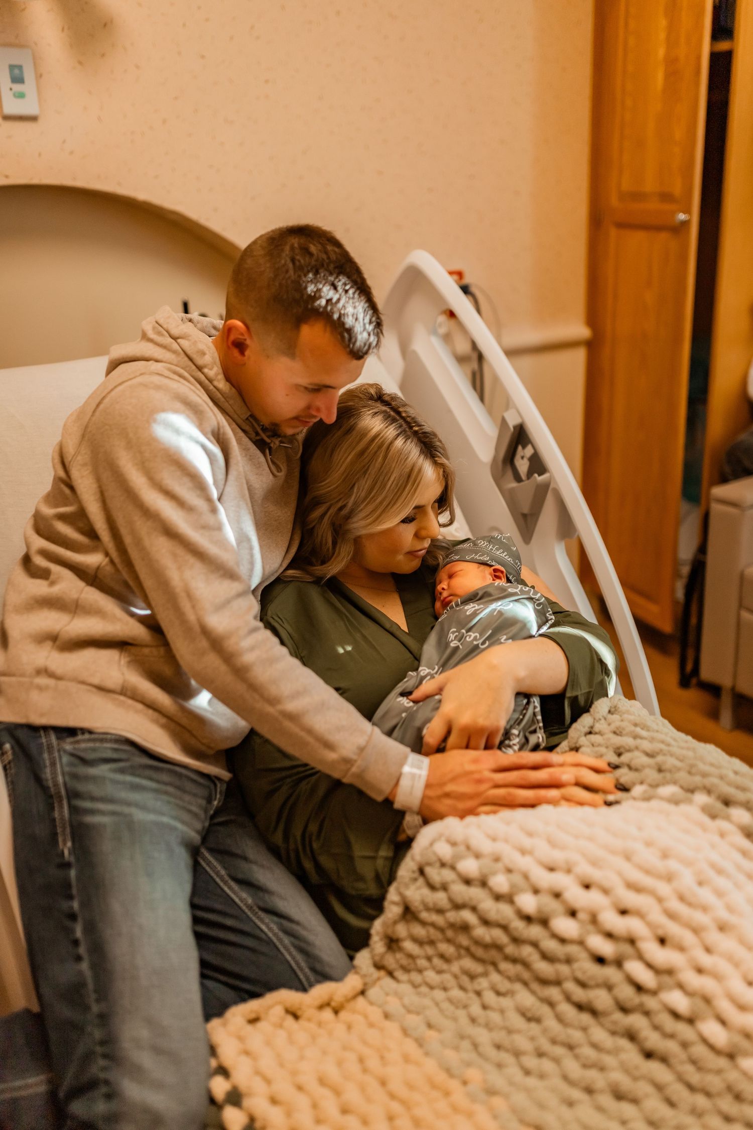 Two parents share an intimate moment with their newborn baby in a hospital room, draped in a cozy knit blanket.