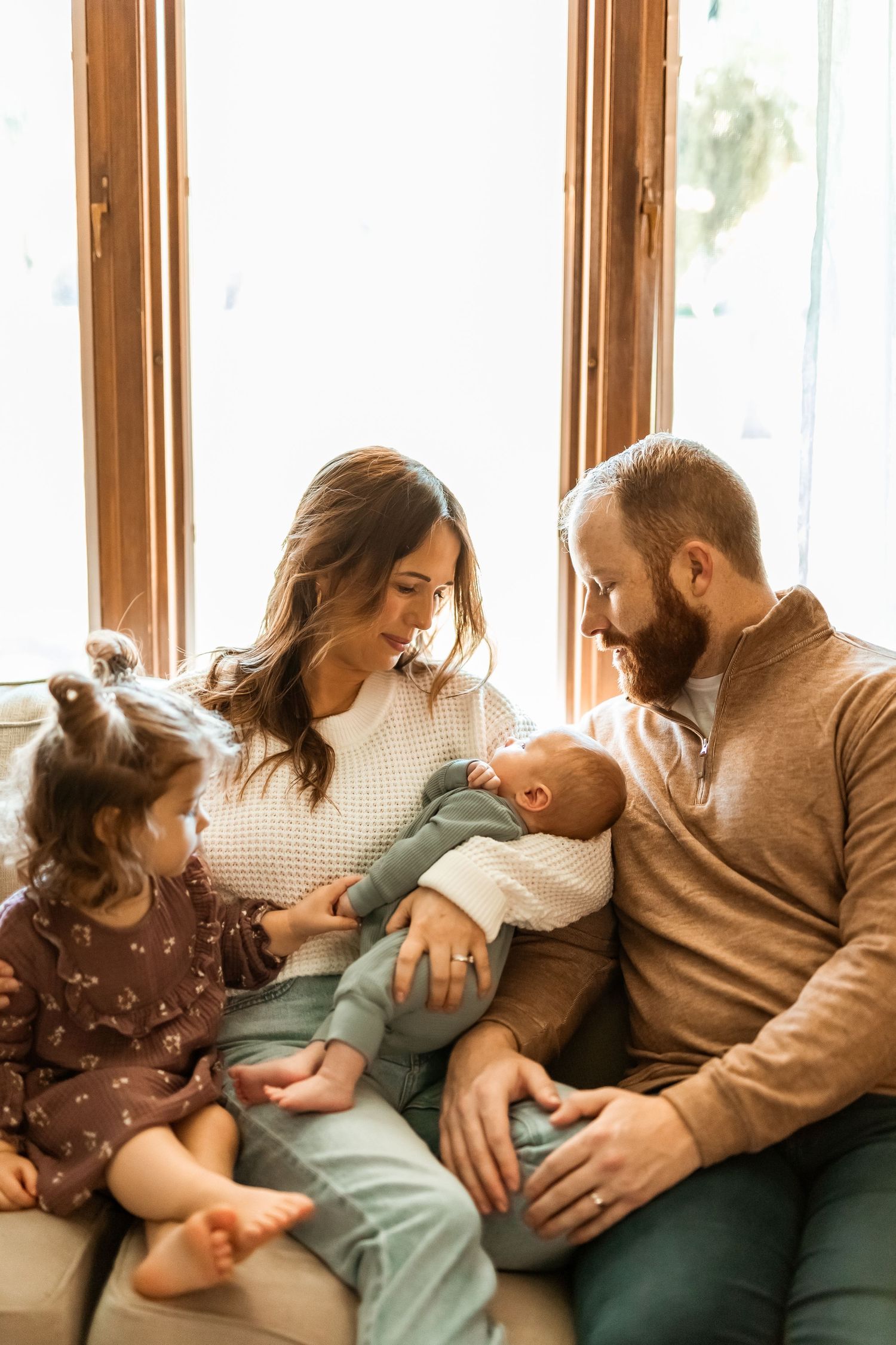 A family sits together on a couch by a window, lovingly gazing at their newborn baby while their toddler looks on.