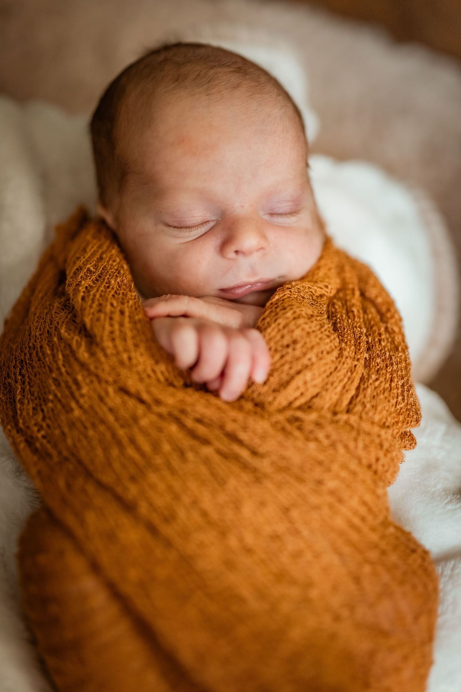 A sleeping newborn peacefully wrapped in a soft golden orange knit blanket against a cream background.