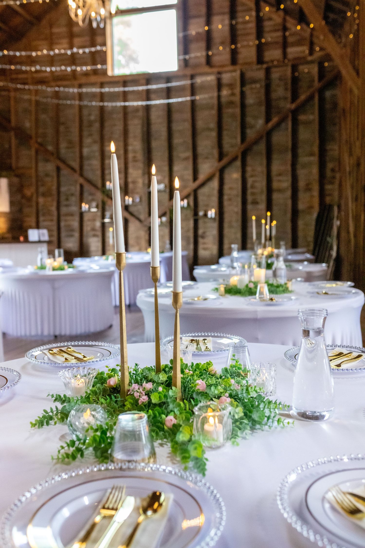 Elegant barn wedding reception table with tall gold candles, greenery centerpiece, and gold cutlery place settings.