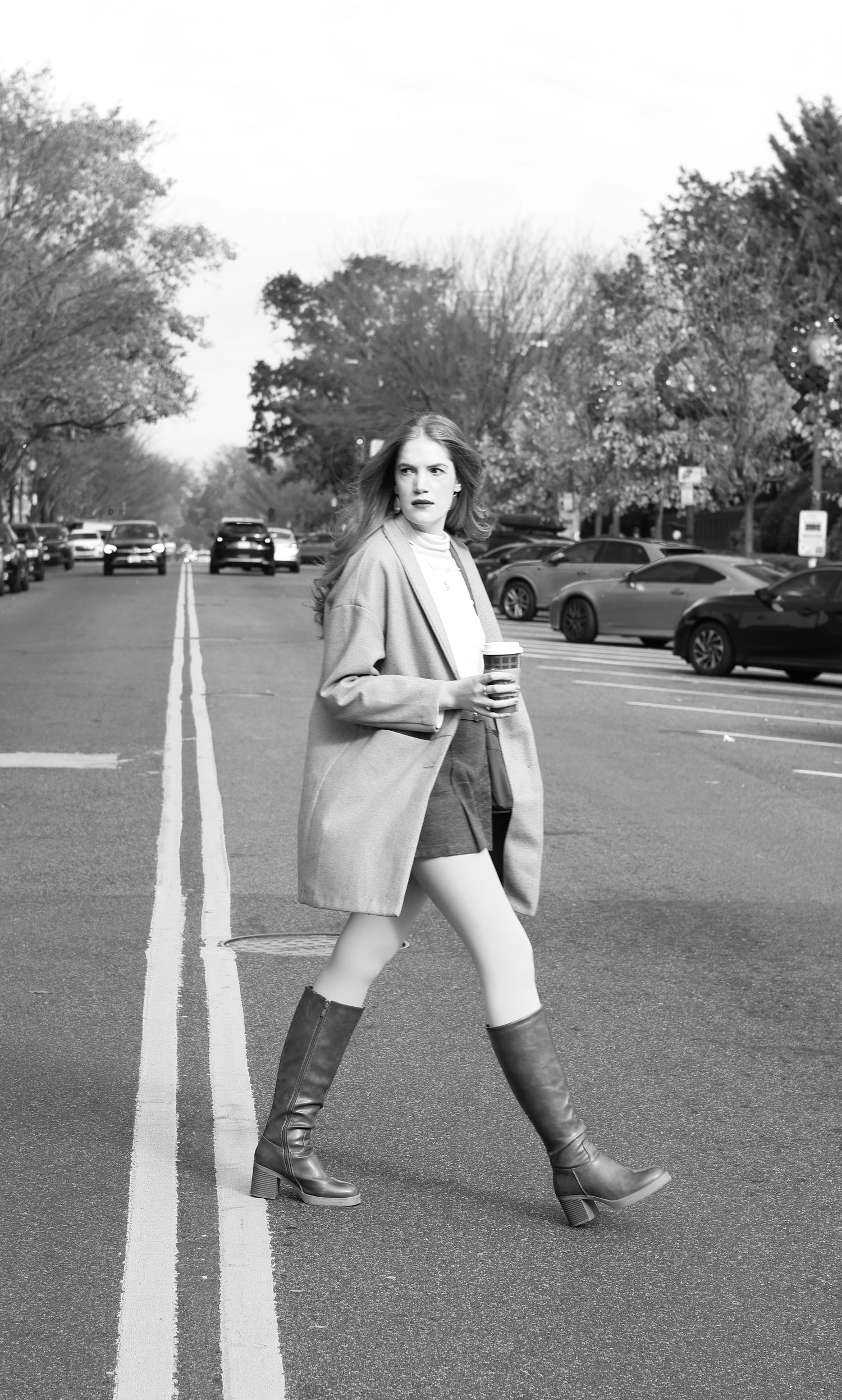 Woman in coat, shorts, and tall boots walks across street holding coffee cup in black and white.
