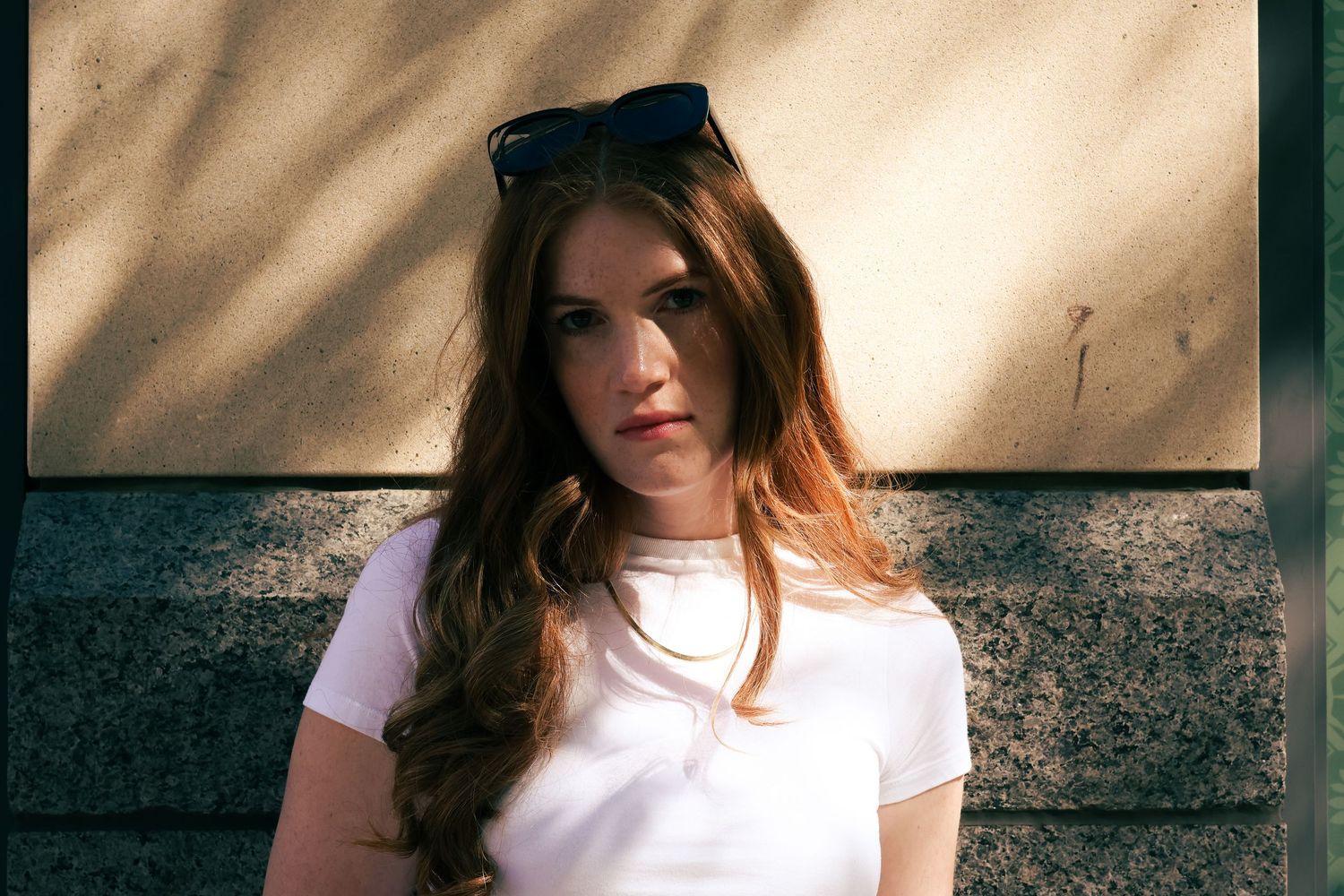 Young woman with long auburn hair and sunglasses on head wearing white t-shirt, posing outdoors near a wall.