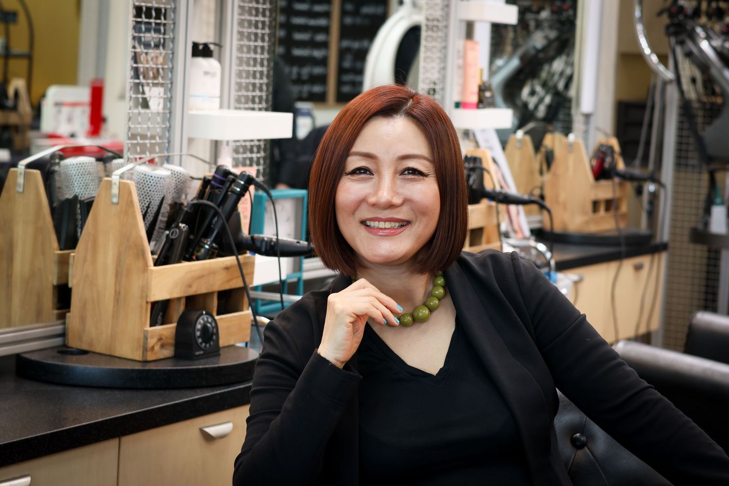 A customer sits at a salon counter with a cheerful expression while resting their chin on their hand.