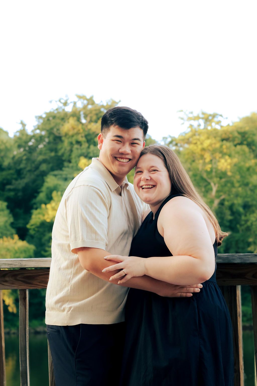 A smiling couple embraces on a wooden deck railing, with lush green trees and calm water glowing in soft golden-hour light behind them.