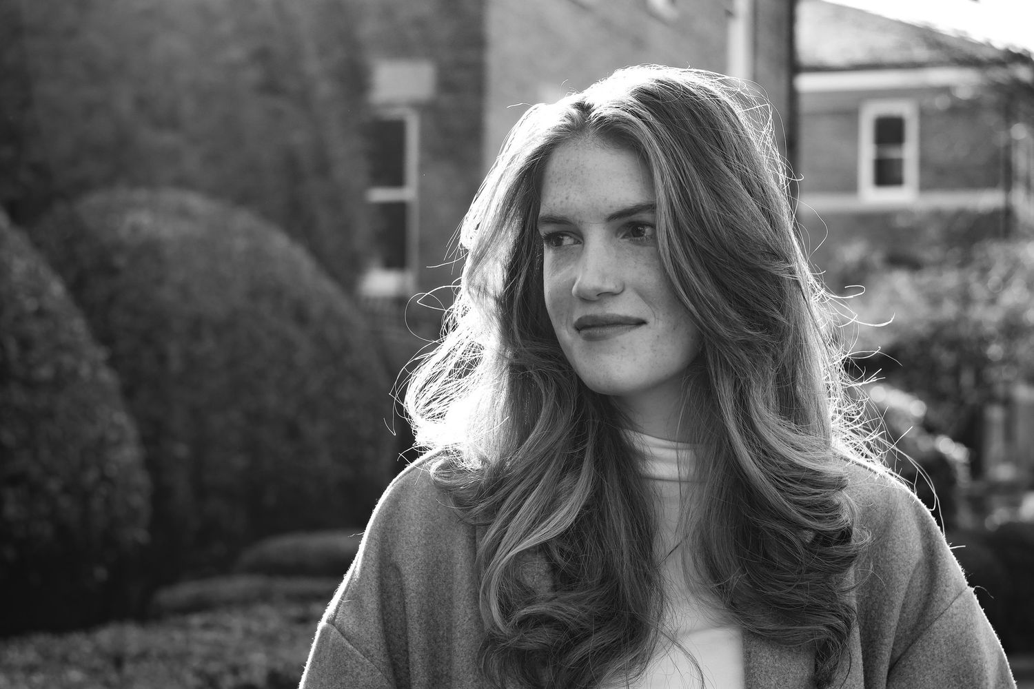 Black and white portrait of a young woman with long wavy hair, posing outdoors near trimmed hedges.