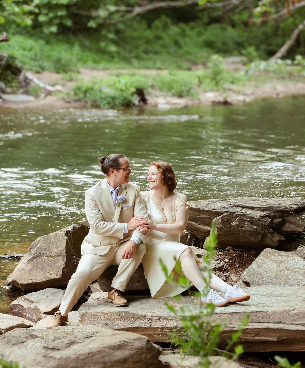 Smiling couple in cream wedding attire sit close together on flat rocks beside a calm, tree-lined river.