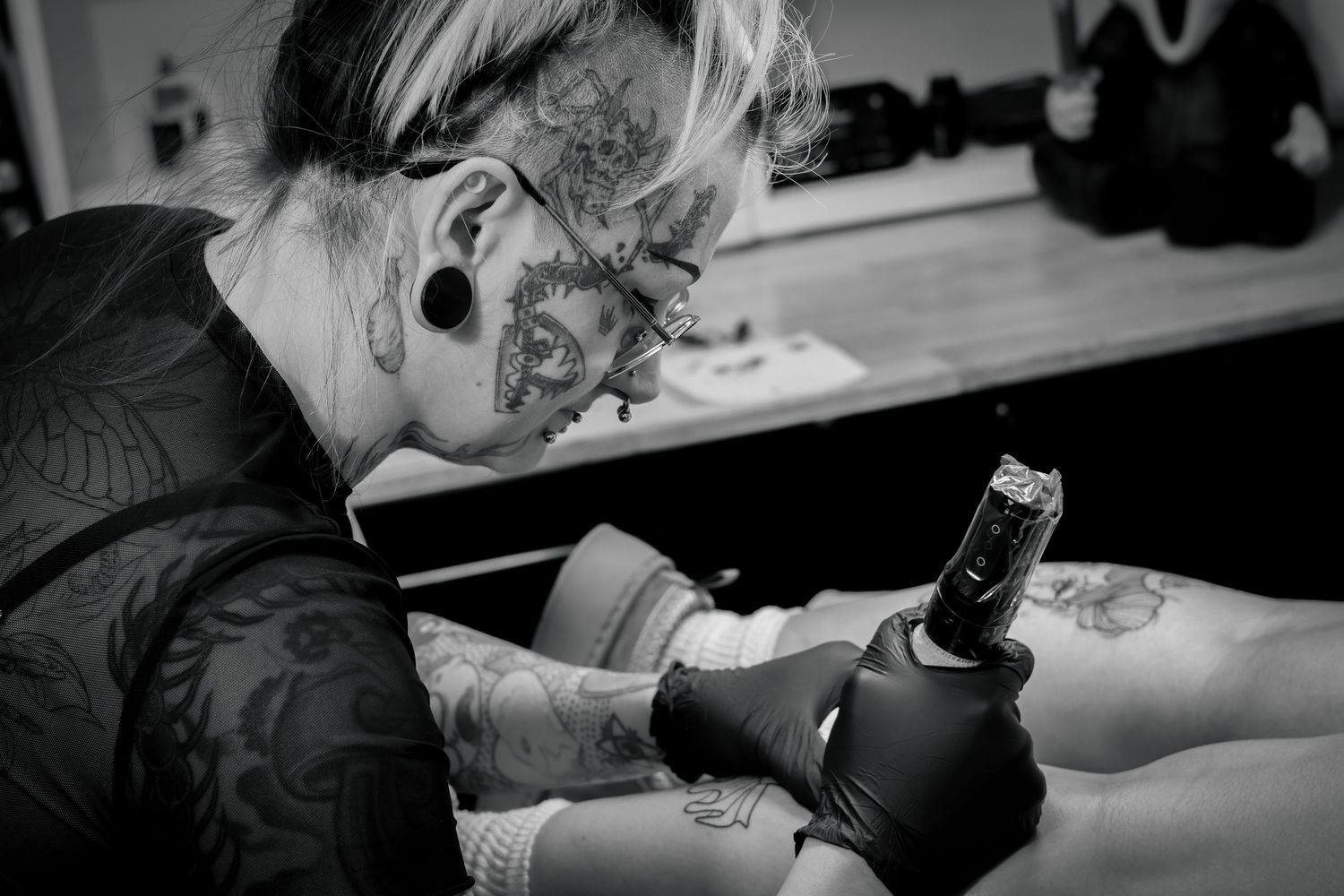 Professional tattoo artist wearing black gloves works on a detailed tattoo design in a studio setting.