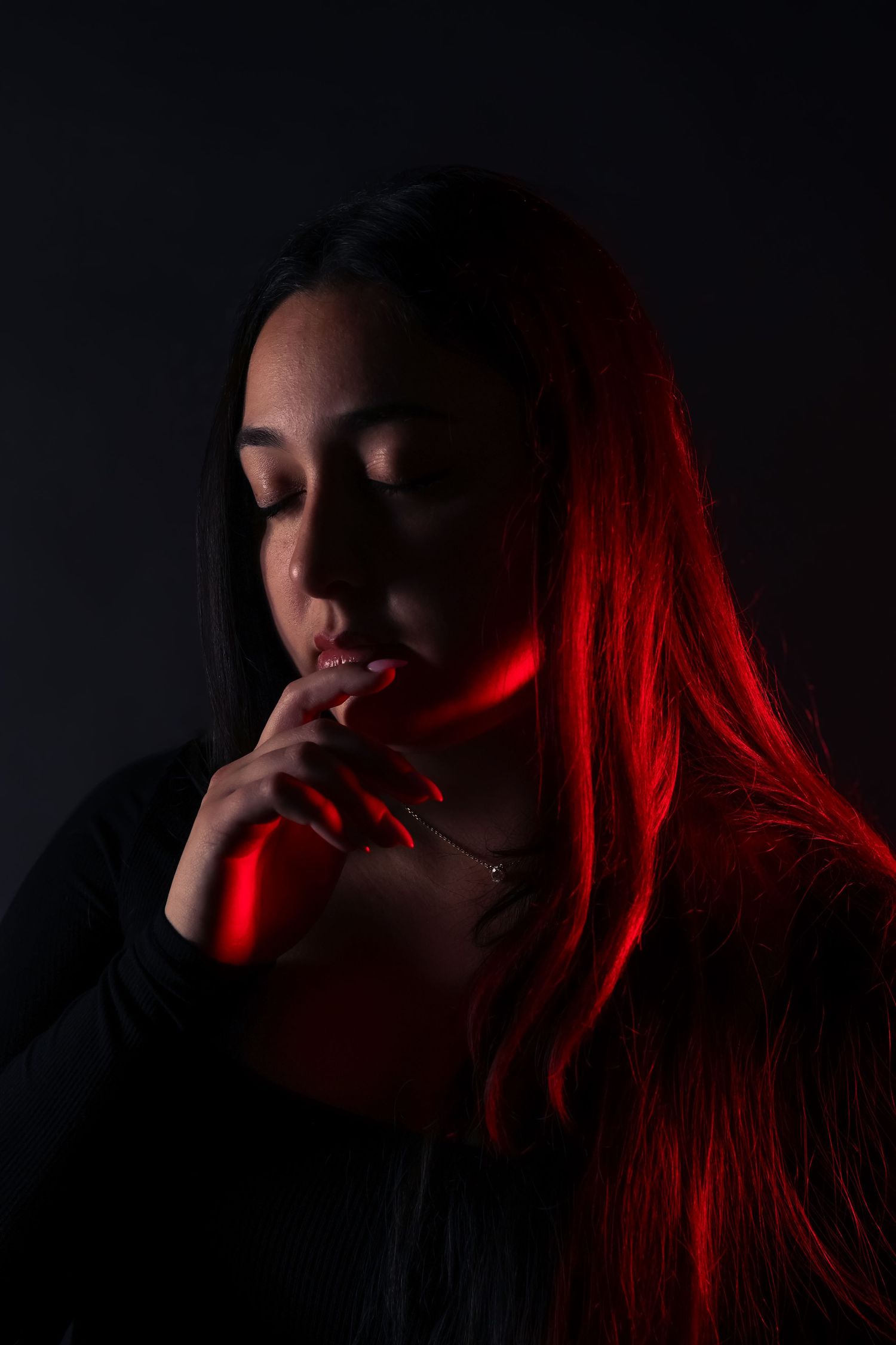 Woman with long hair illuminated by red light, eyes closed, hand near chin, dark background.